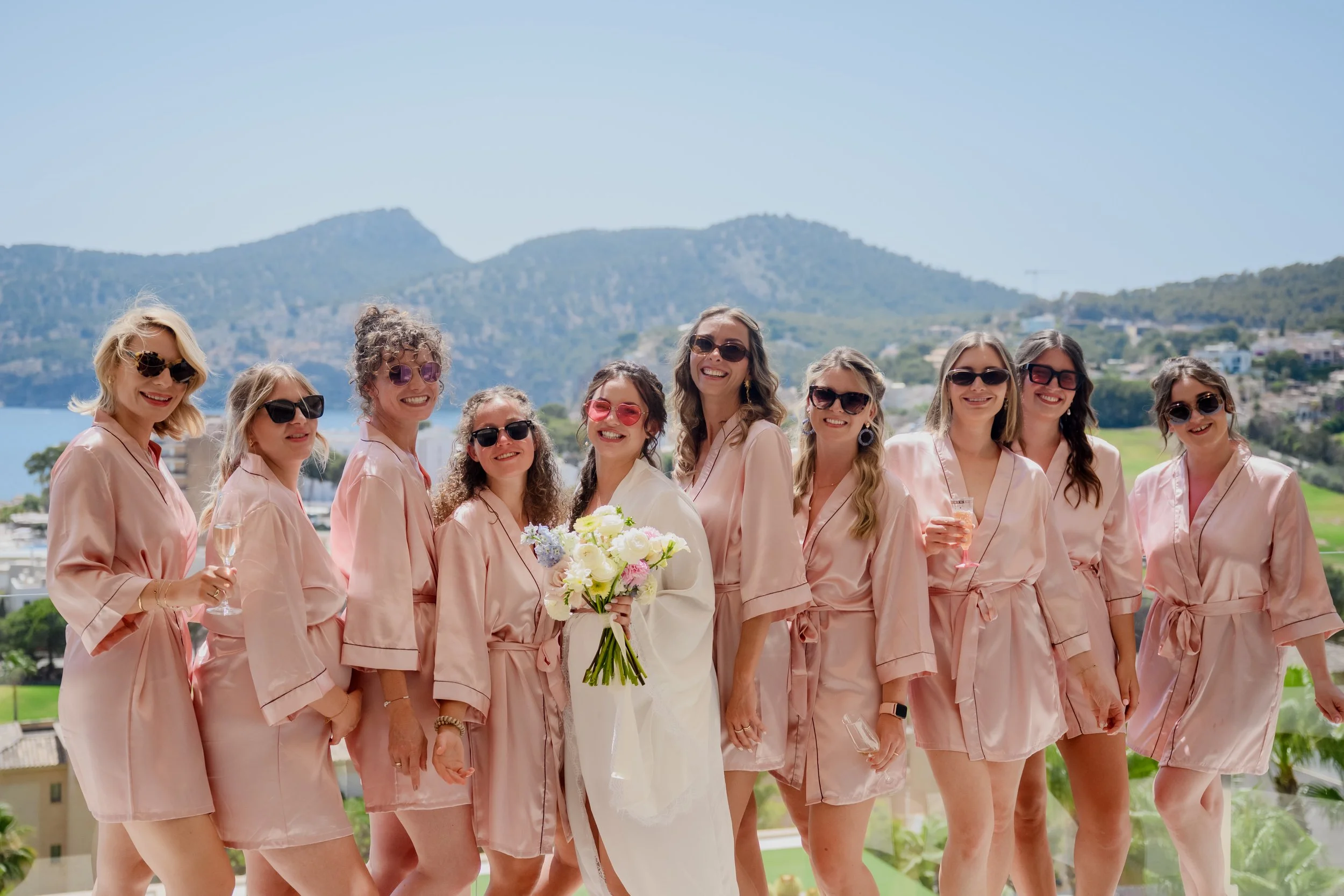 Bride and bridesmaids wearing matching pale pink robes, smiling outdoors with mountains in the background, some holding drinks, one holding a bouquet.