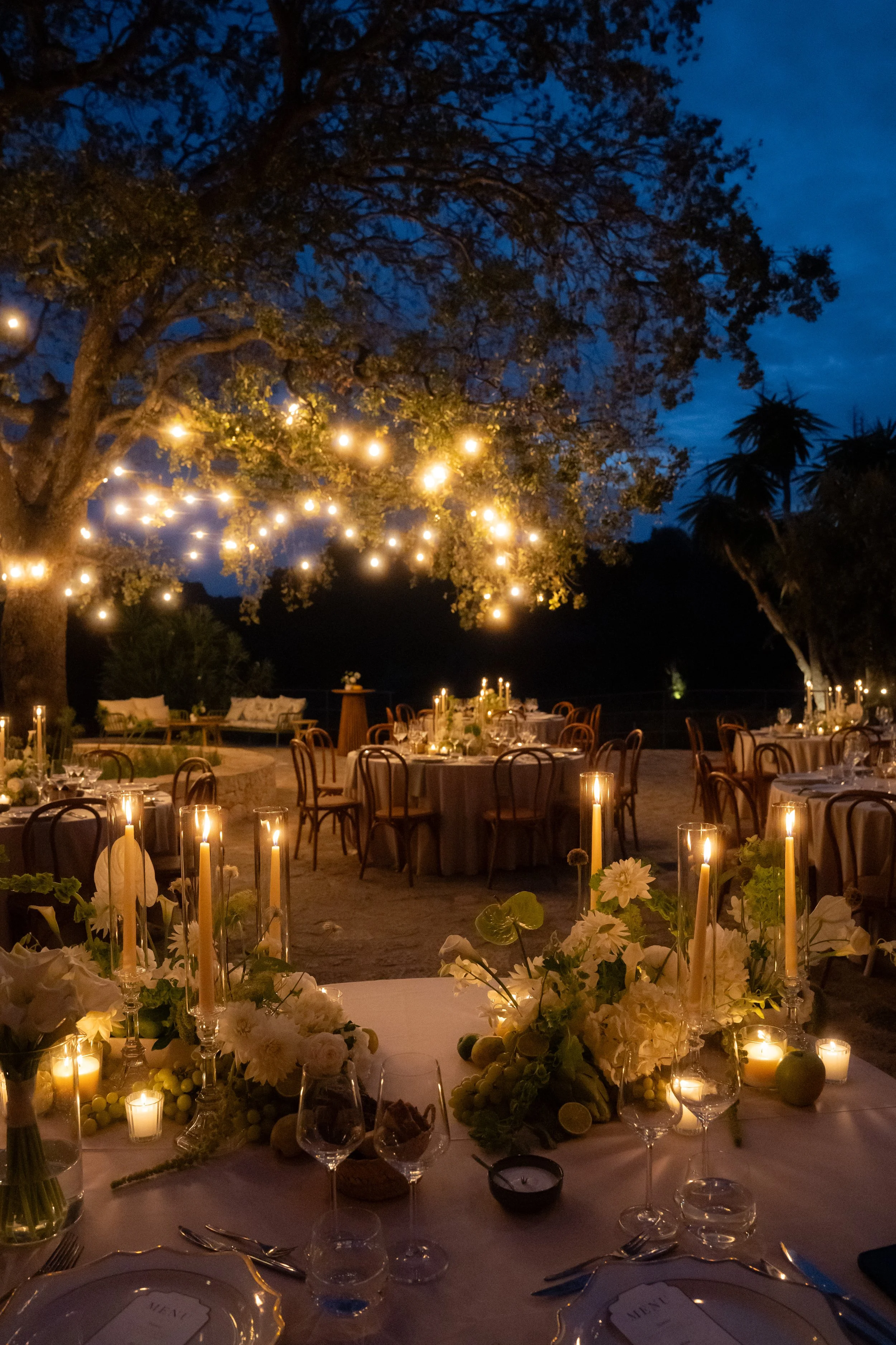 Outdoor wedding reception at night with tables decorated with flowers, candles, and string lights hanging from tree branches.