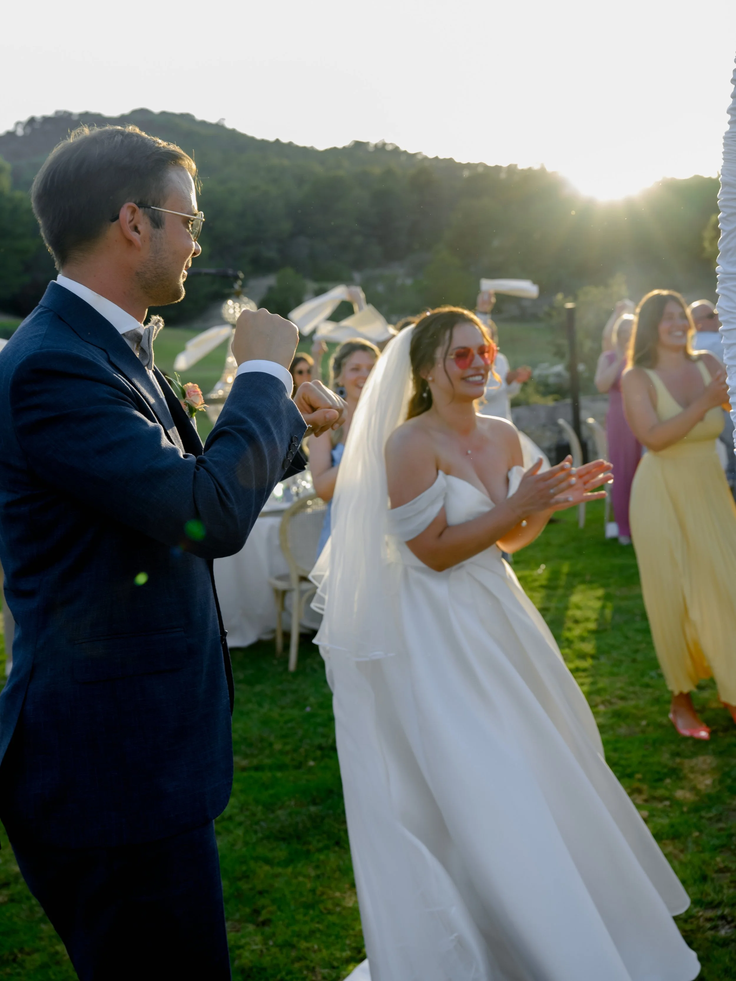 A wedding couple dancing outdoors at sunset, with guests clapping and smiling in the background.