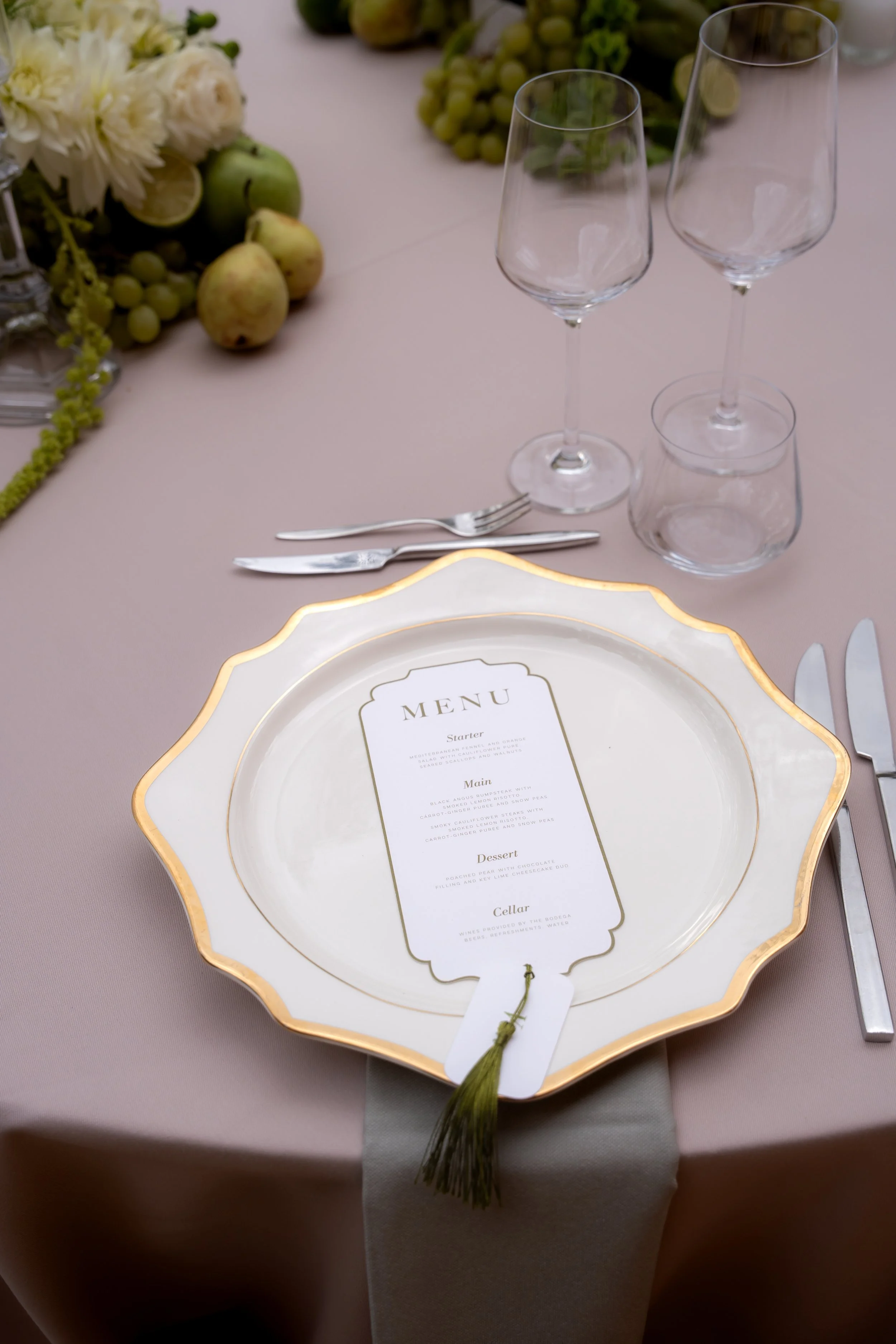 A formal table setting with a decorative white and gold plate, a menu card, silverware, empty glasses, and a centerpiece of flowers and fruit on a pink tablecloth.