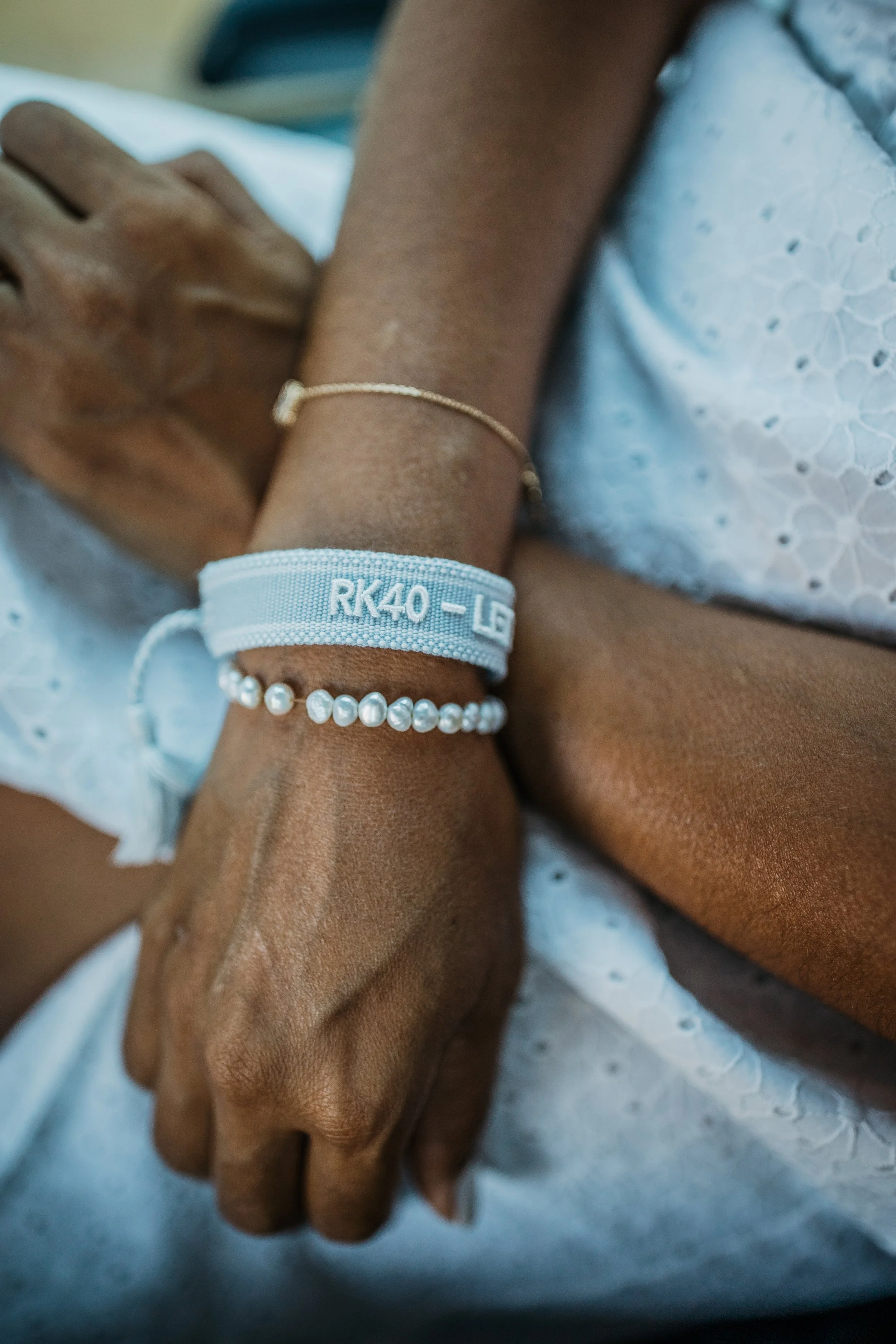 Close-up of a person's hand and wrist resting on their lap, wearing a pearl bracelet, a fabric wristband with text, and a delicate gold bracelet, with the person wearing a white textured fabric garment.