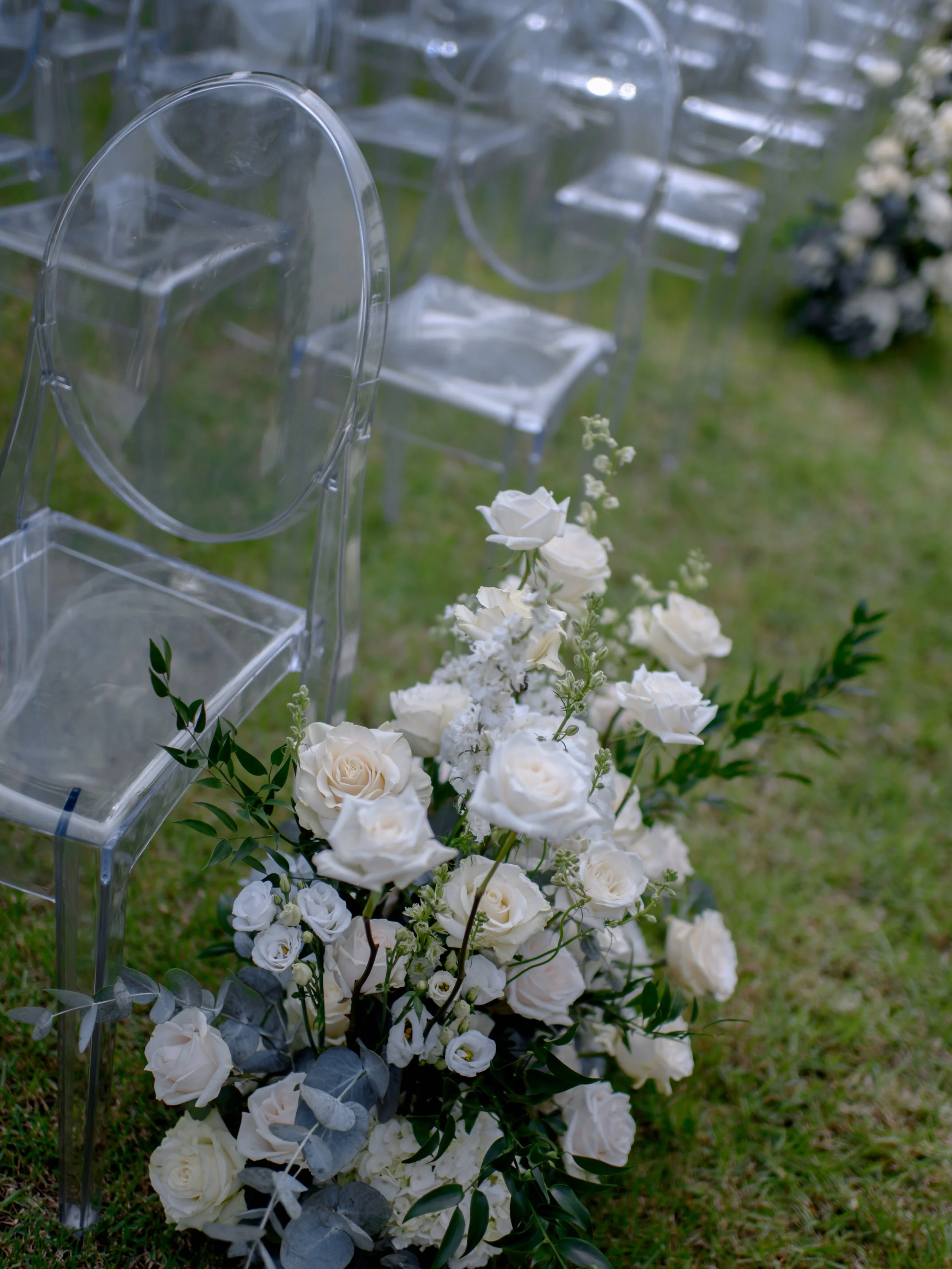 Clear acrylic chairs and a floral arrangement of white roses and greenery on a grassy area, likely outdoors for a wedding or event.