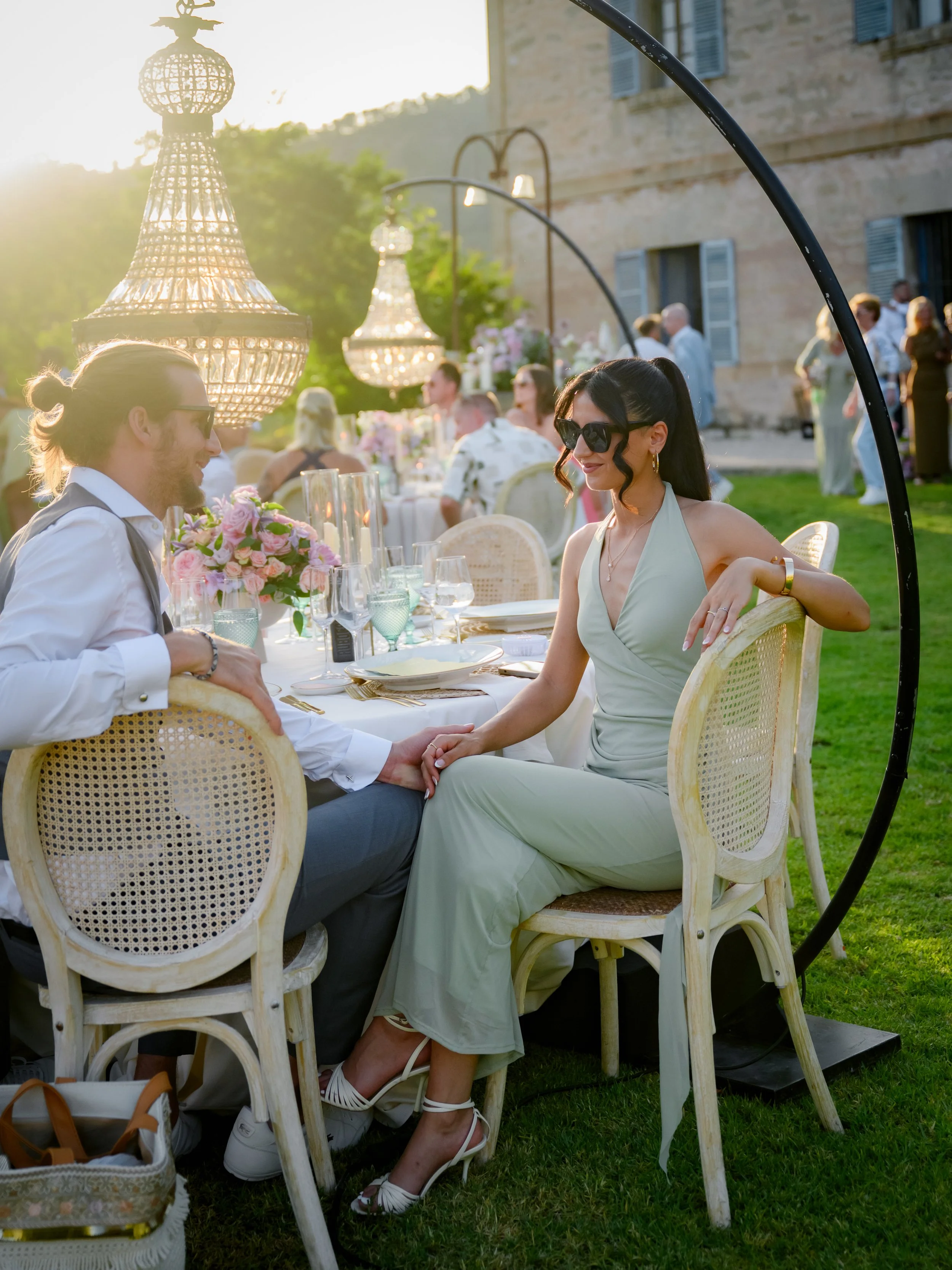 A couple sitting outdoors at a table during a wedding or fancy event, holding hands and smiling. The woman is wearing a light green dress and sunglasses, and the man has long hair tied back, wearing a white shirt and dark vest. The table is decorated