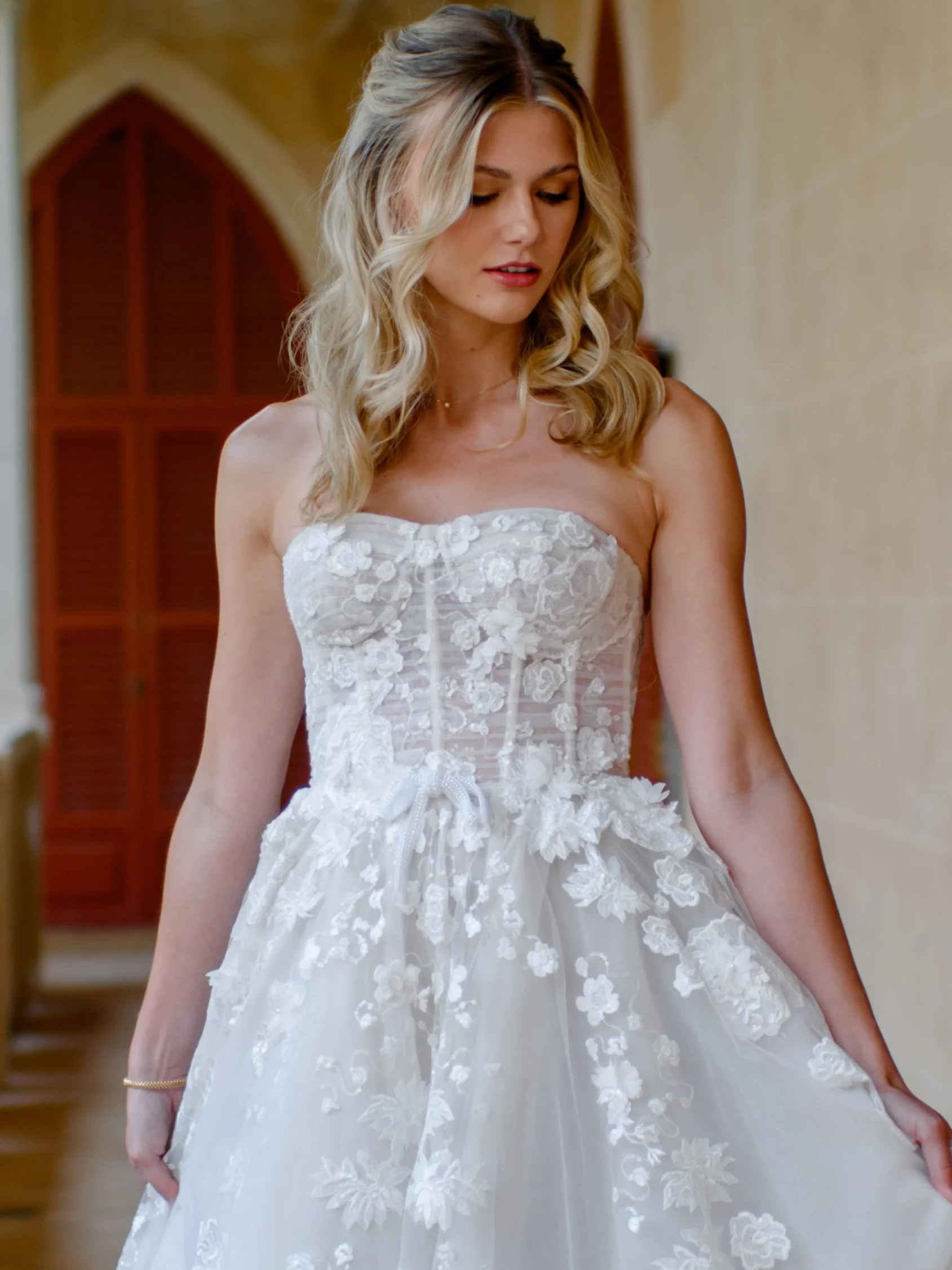A woman trying on a strapless white wedding dress with lace details in a fitting room.