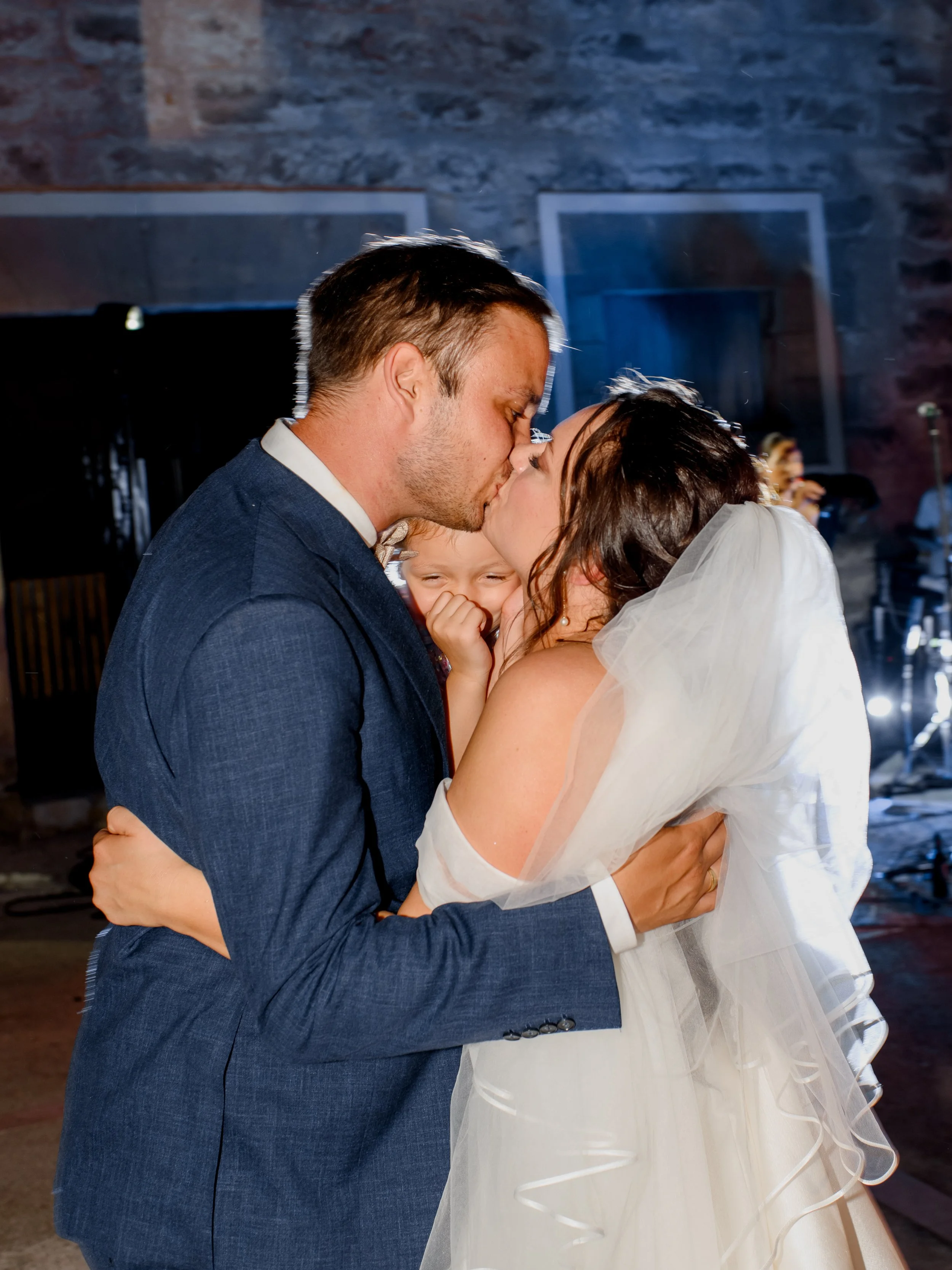 A groom and bride sharing a kiss at their wedding, with a young girl smiling in the background, indoors with stone walls and celebratory lighting.