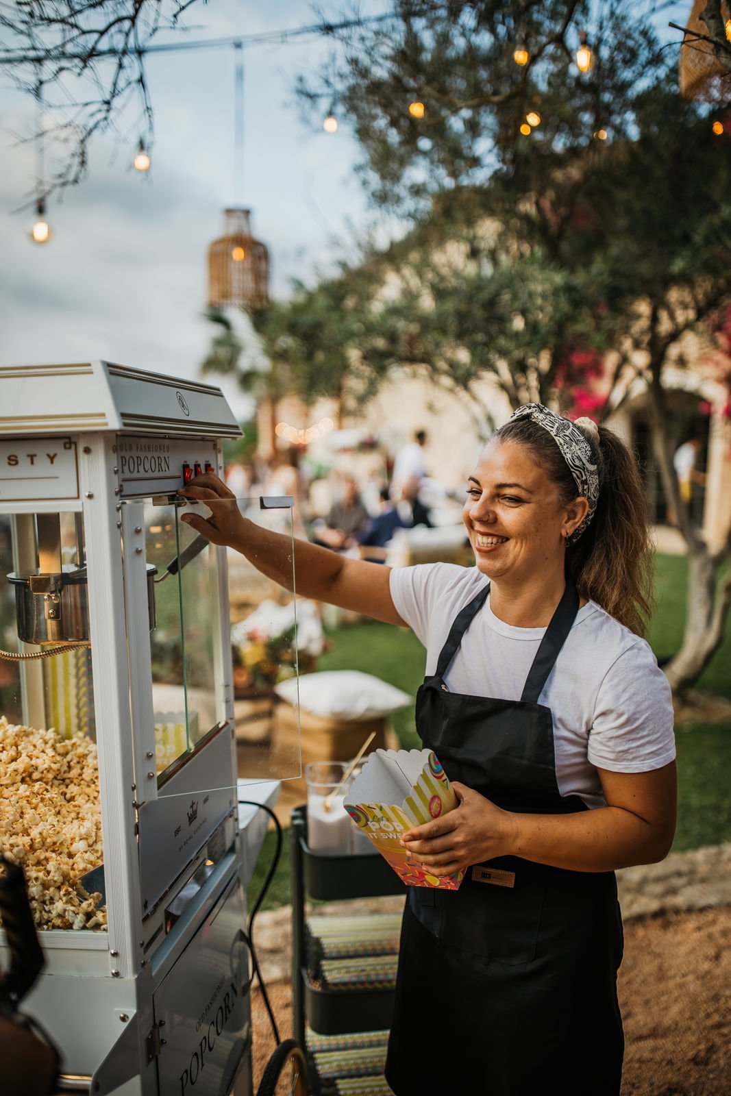 A woman in a white t-shirt and black apron smiling while operating a popcorn machine outdoors during evening with string lights hanging above and people in the background.