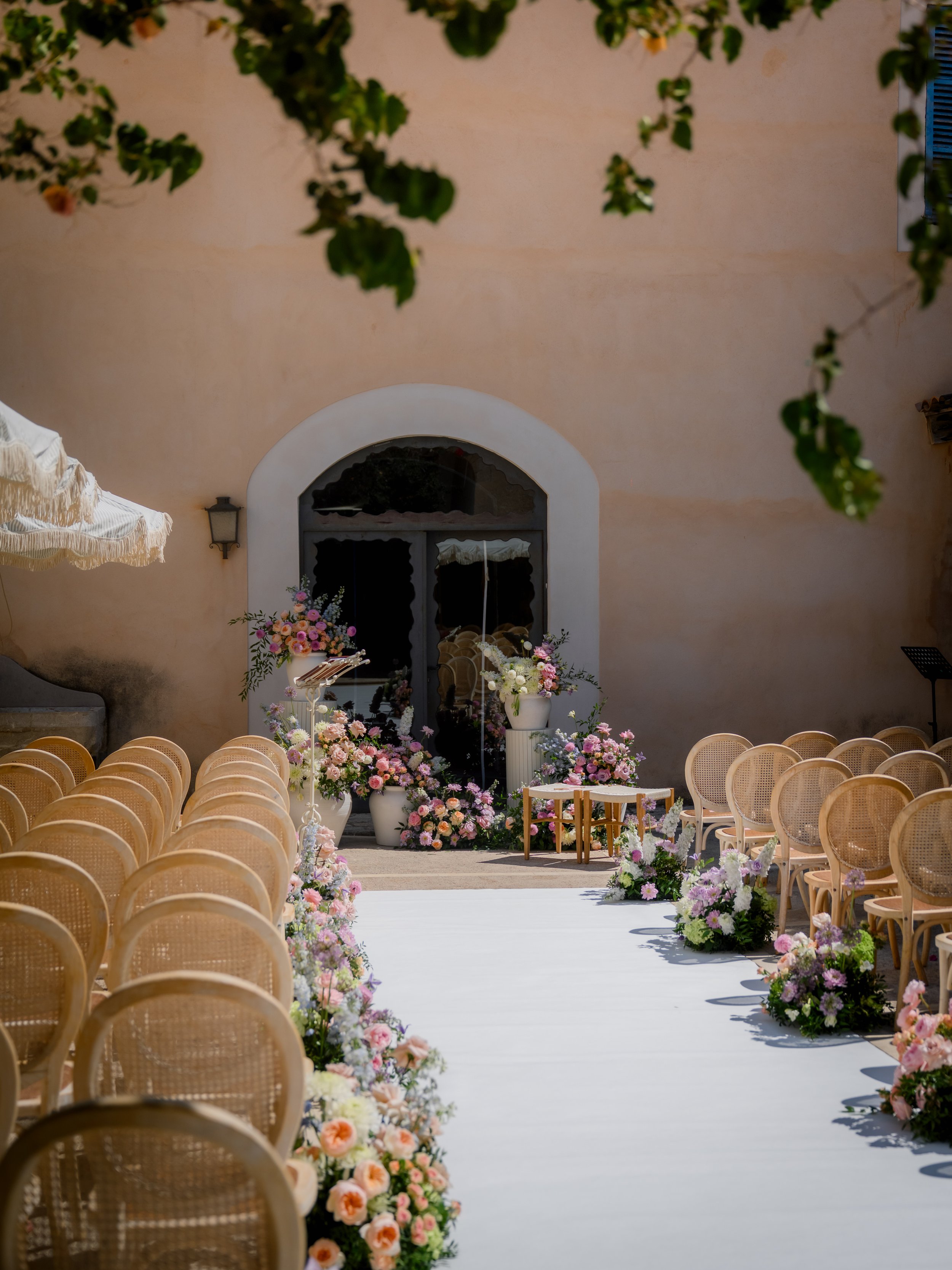 An outdoor wedding ceremony setup with rows of beige chairs, floral arrangements with pink and purple flowers lining the aisle, and a floral altar in front of a beige wall with a window.