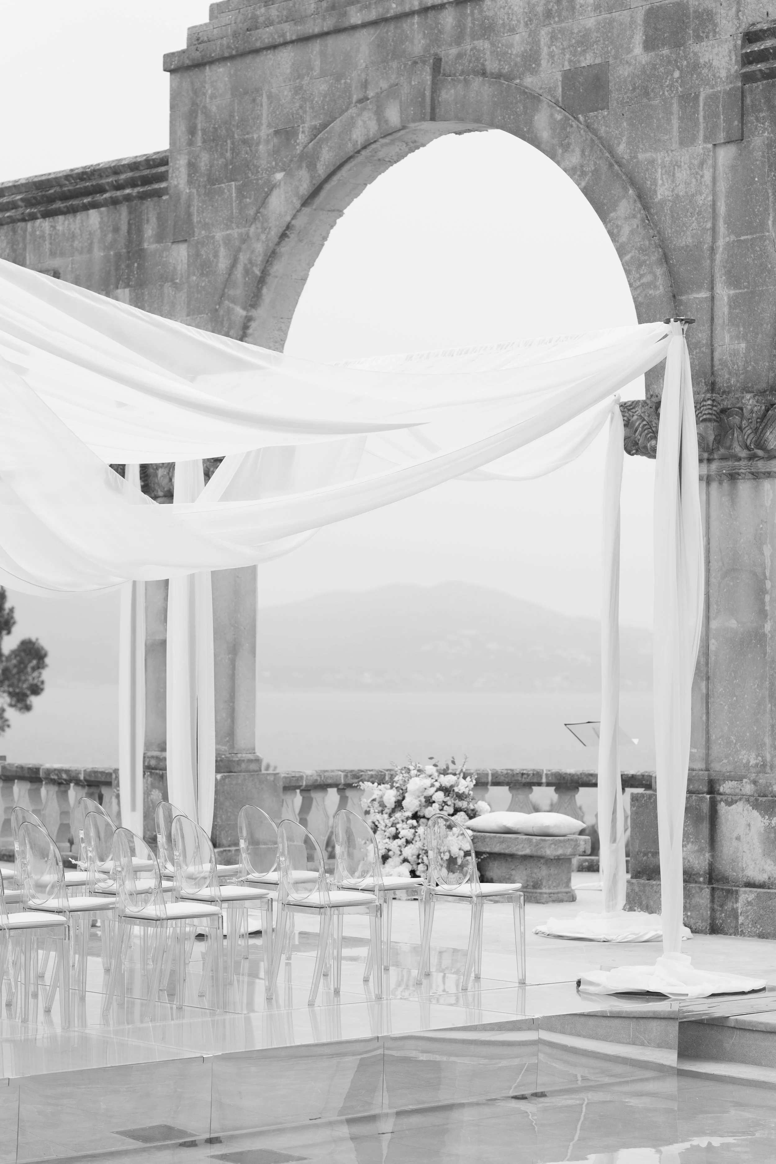 A black and white image of an outdoor wedding altar with sheer fabric drapes, transparent chairs, a floral arrangement, and a stone arch, with a scenic backdrop of mountains and a body of water.