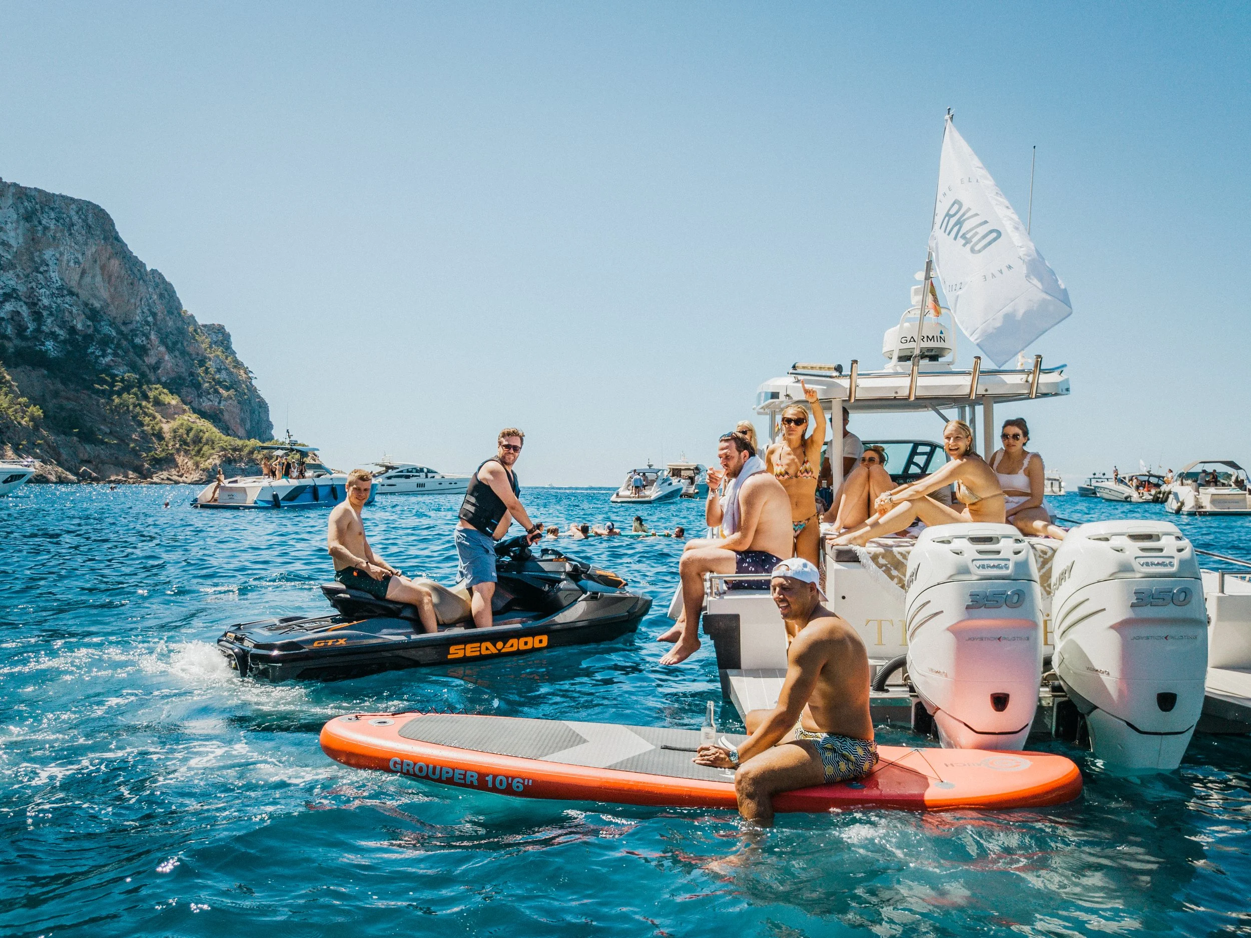 Group of people enjoying a day on boats and jet skis in the ocean near a rocky coastline.