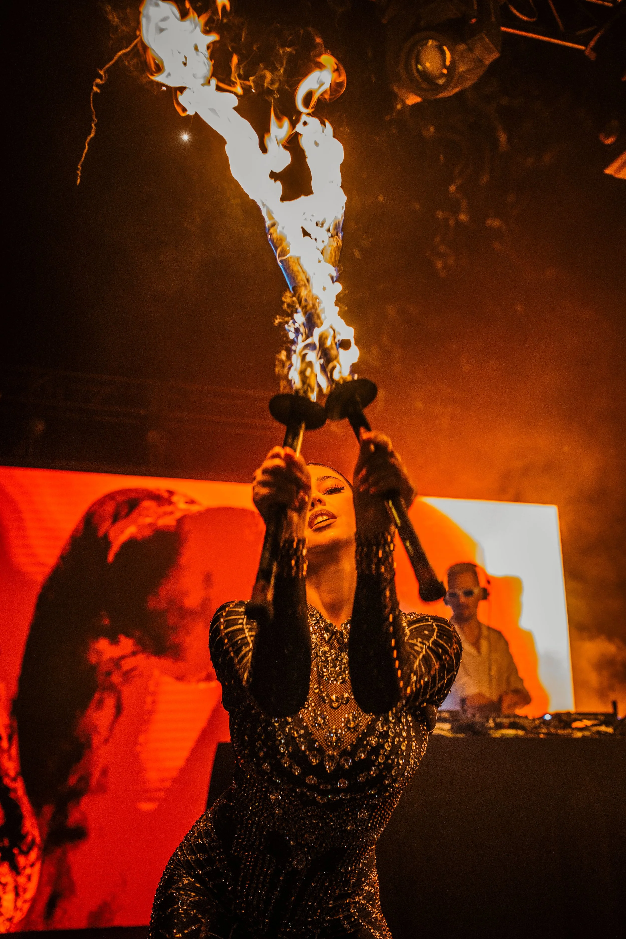 A performer holding two flaming torches above her head on stage, with a background screen showing an artist in a black and gold outfit and a DJ in the background.