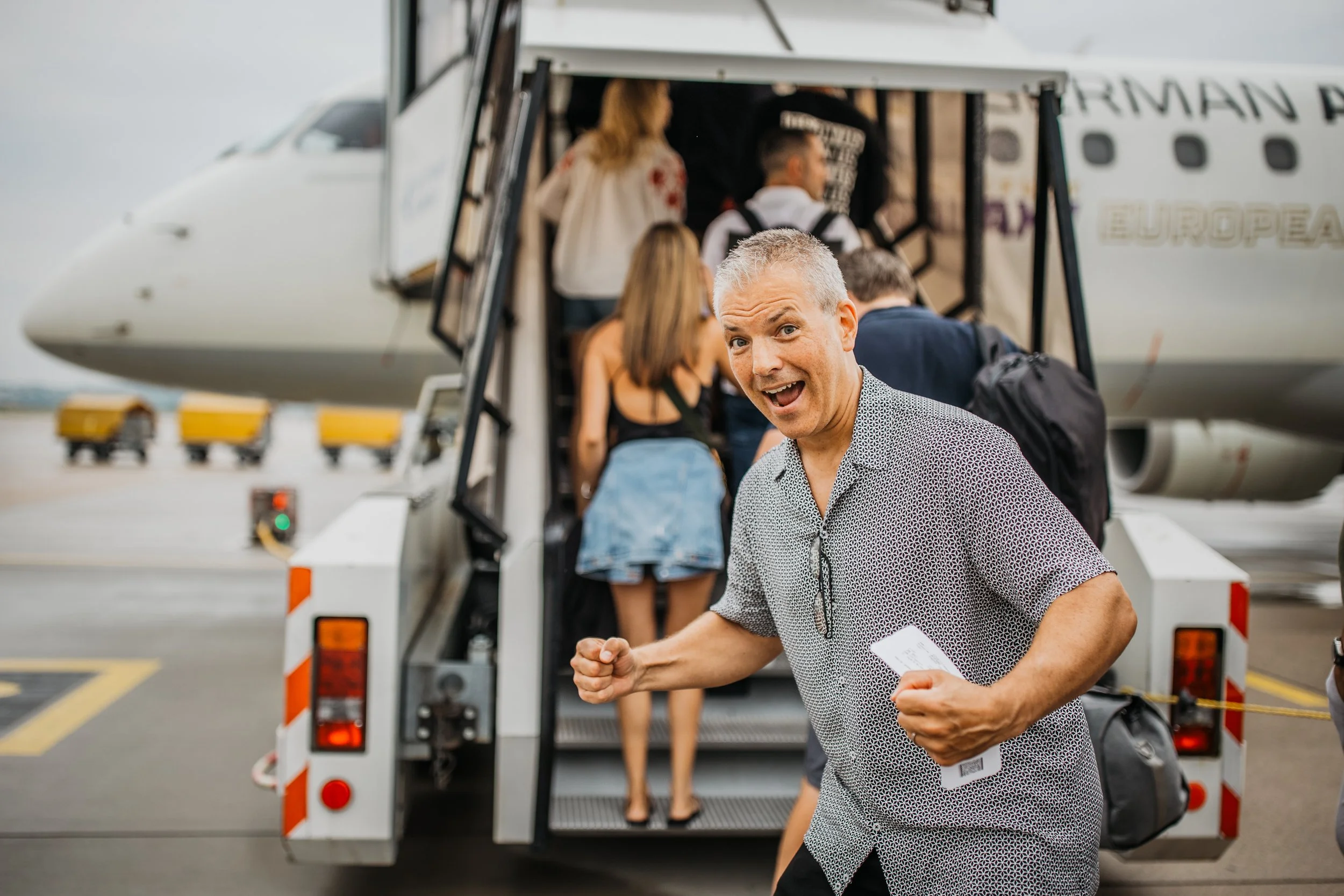 A man with glasses hanging on his shirt, smiling with enthusiasm, standing in front of an airplane boarding stairs at an airport, with other travelers in line boarding the aircraft.