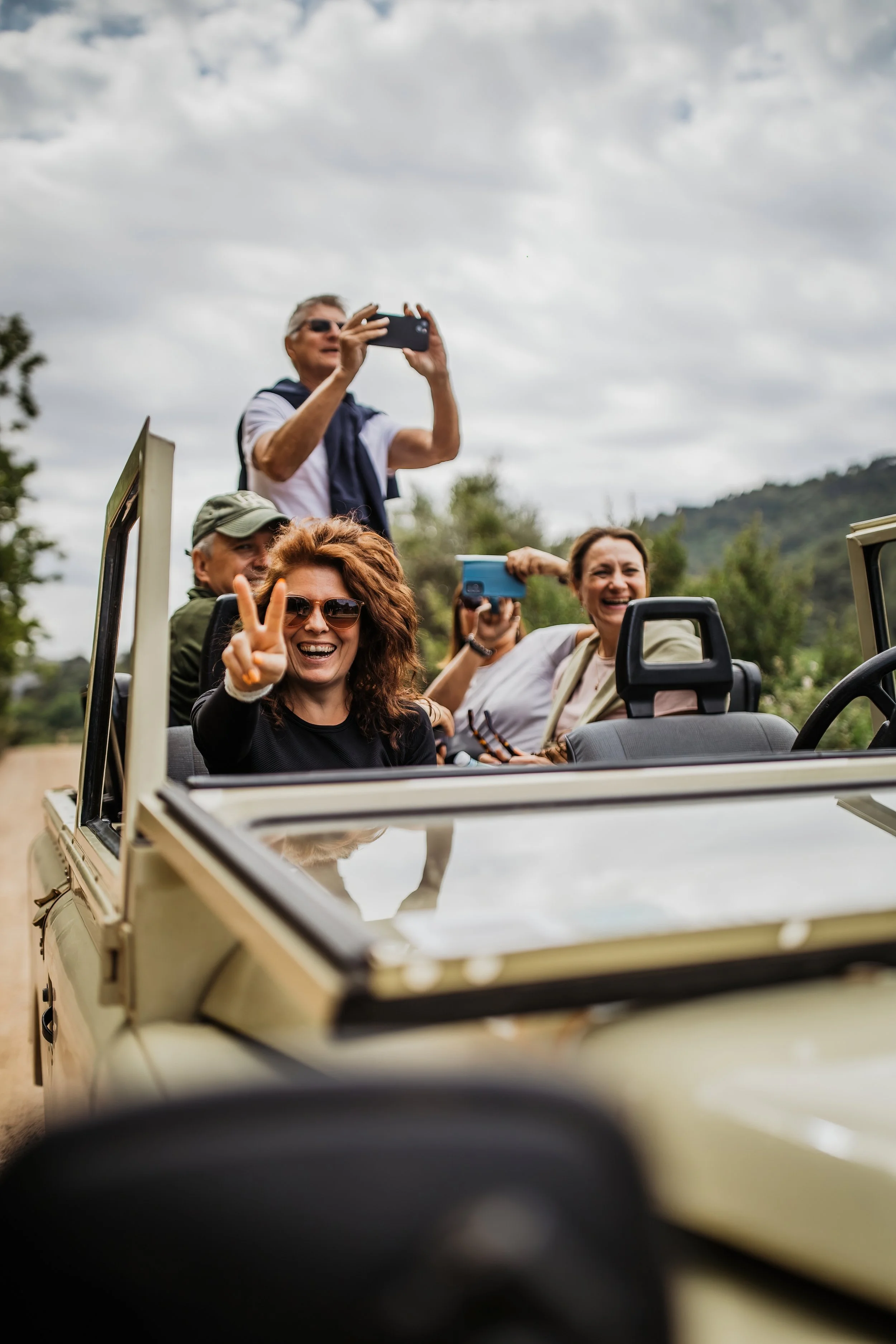 A group of friends enjoying a safari ride in an open-top vehicle outdoors, with some taking photos and smiling.