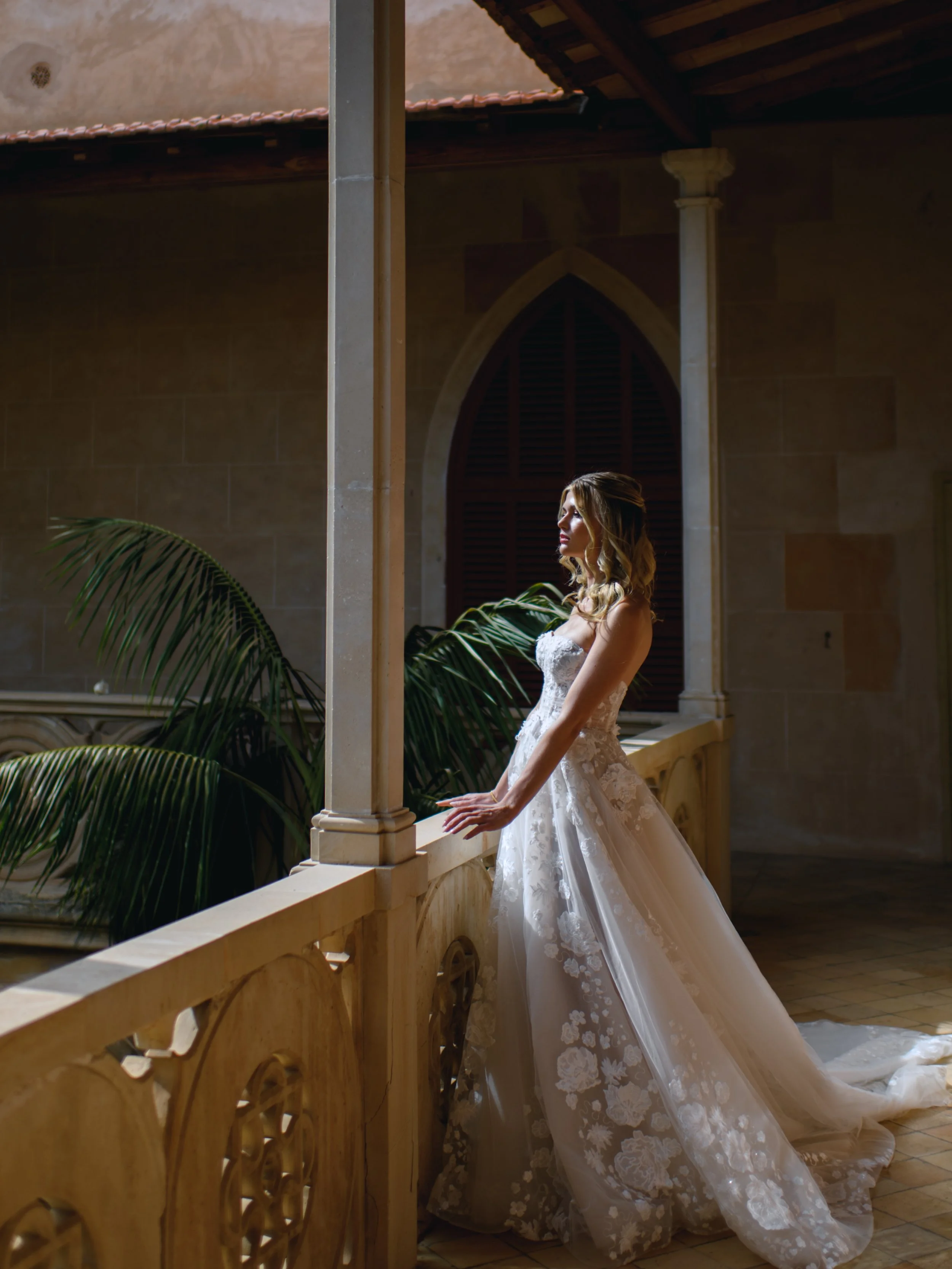A bride in a white wedding dress standing on a balcony with her hands resting on a stone railing. She is looking to the side with her eyes closed, in a dimly lit, elegant interior space with architectural details and lush green plants in the backgrou