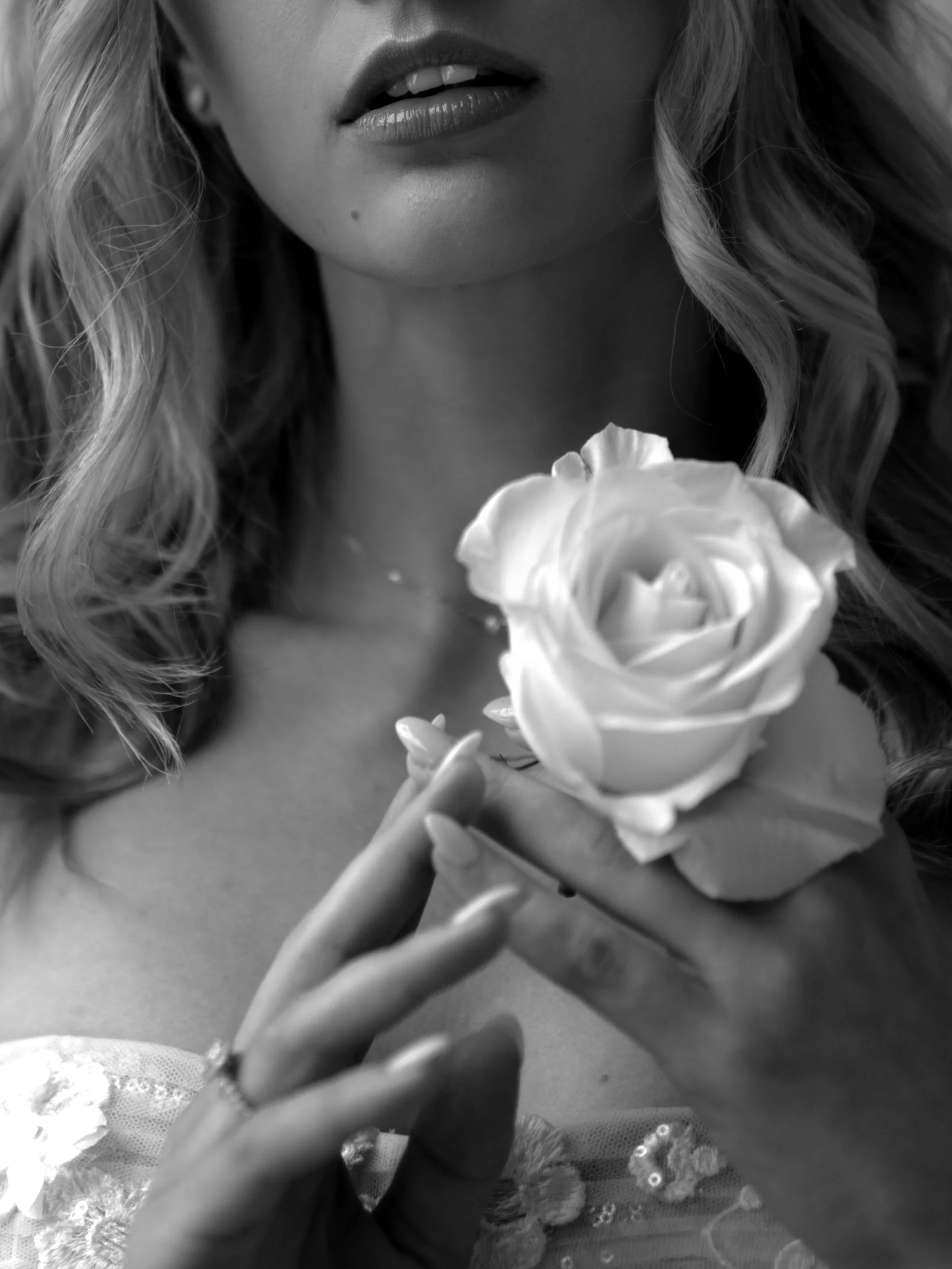Close-up black and white photo of a woman holding a white rose near her chest, with her lips slightly parted and a small beauty mark on her face, with wavy hair framing her face.