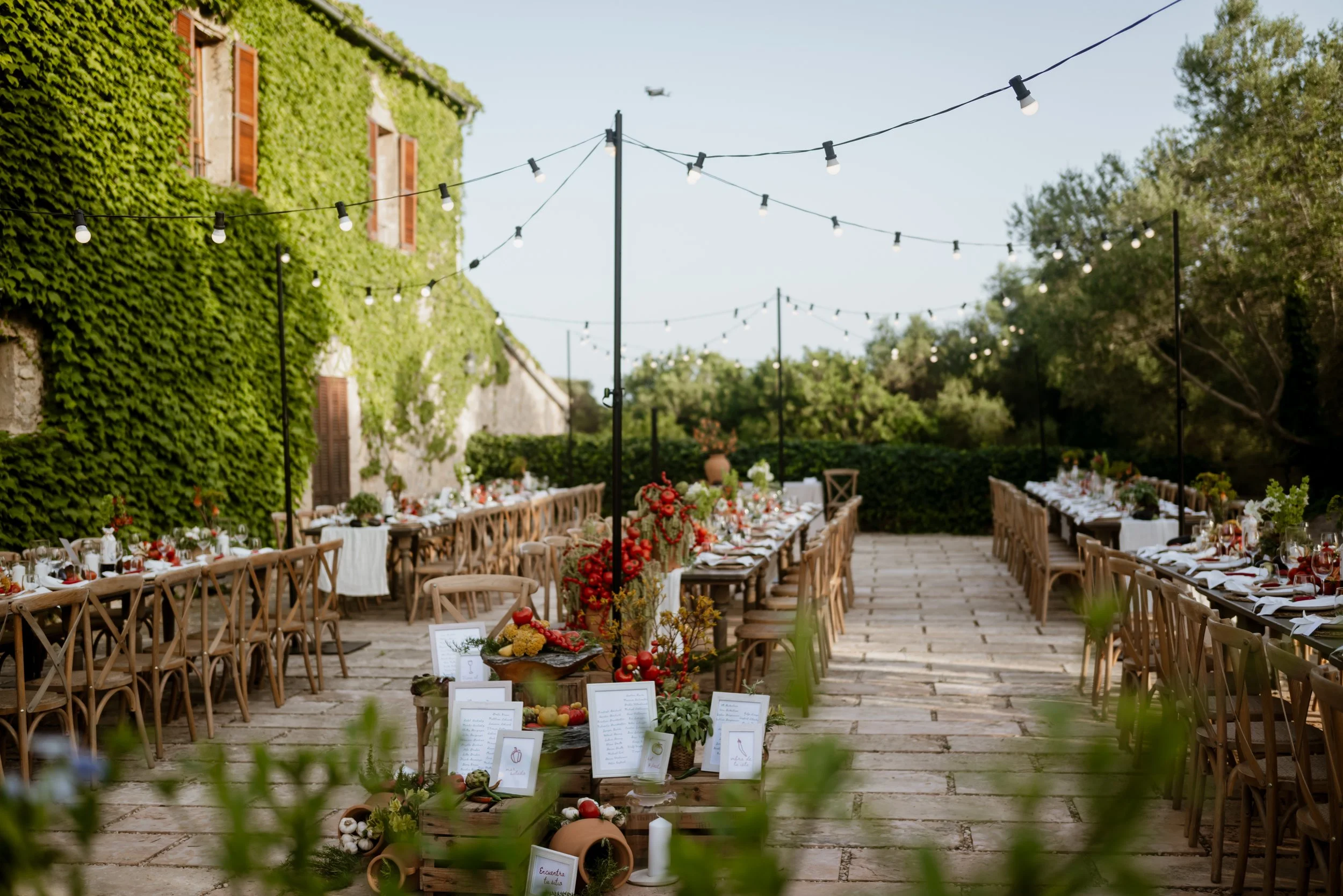 Outdoor dining area with long tables and wooden chairs decorated with flowers, set for a festive event, under string lights with a green ivy-covered building on the left and trees in the background.