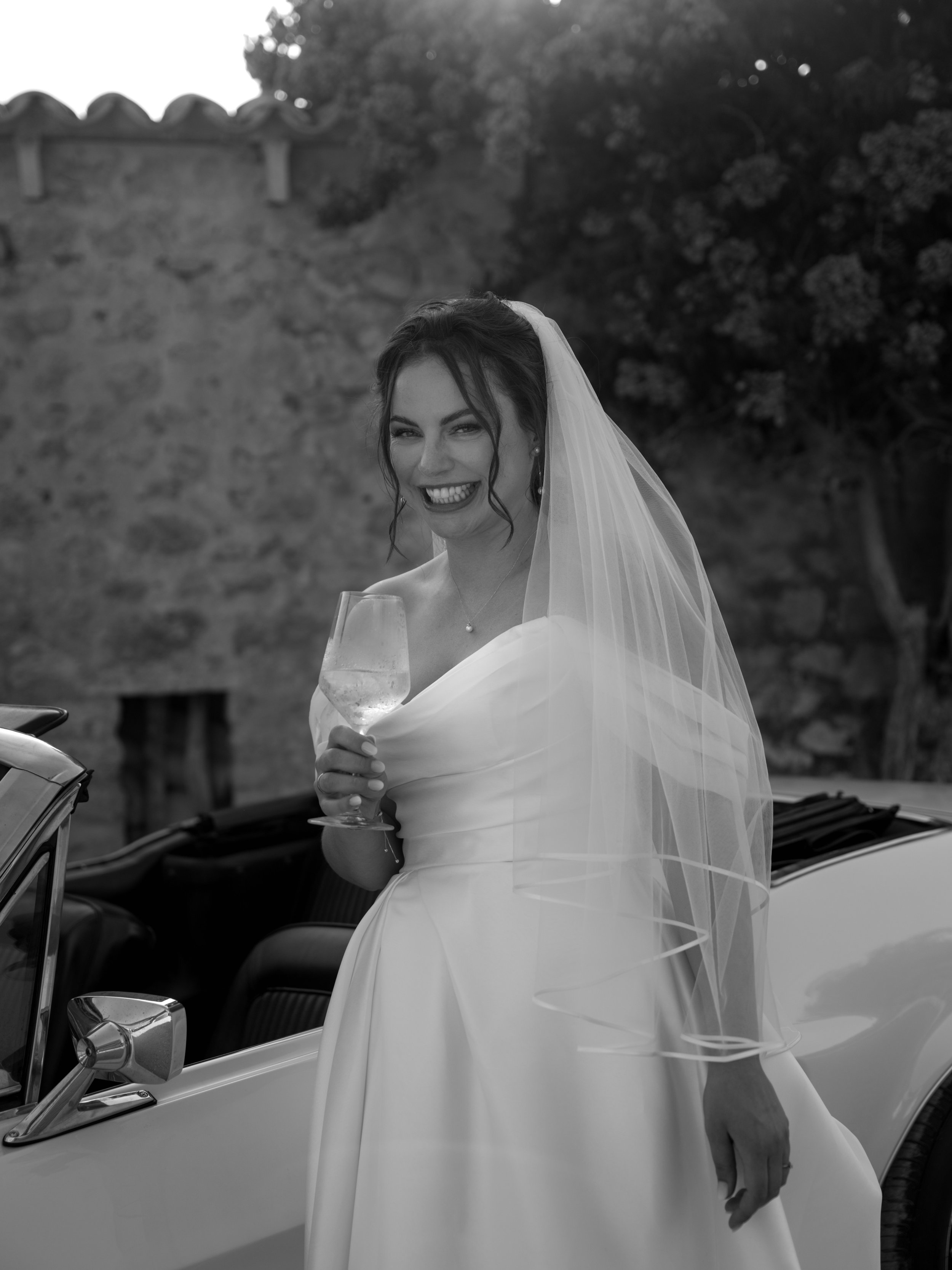 A smiling bride in a wedding dress and veil, holding a glass of wine, standing next to a convertible car outdoors.