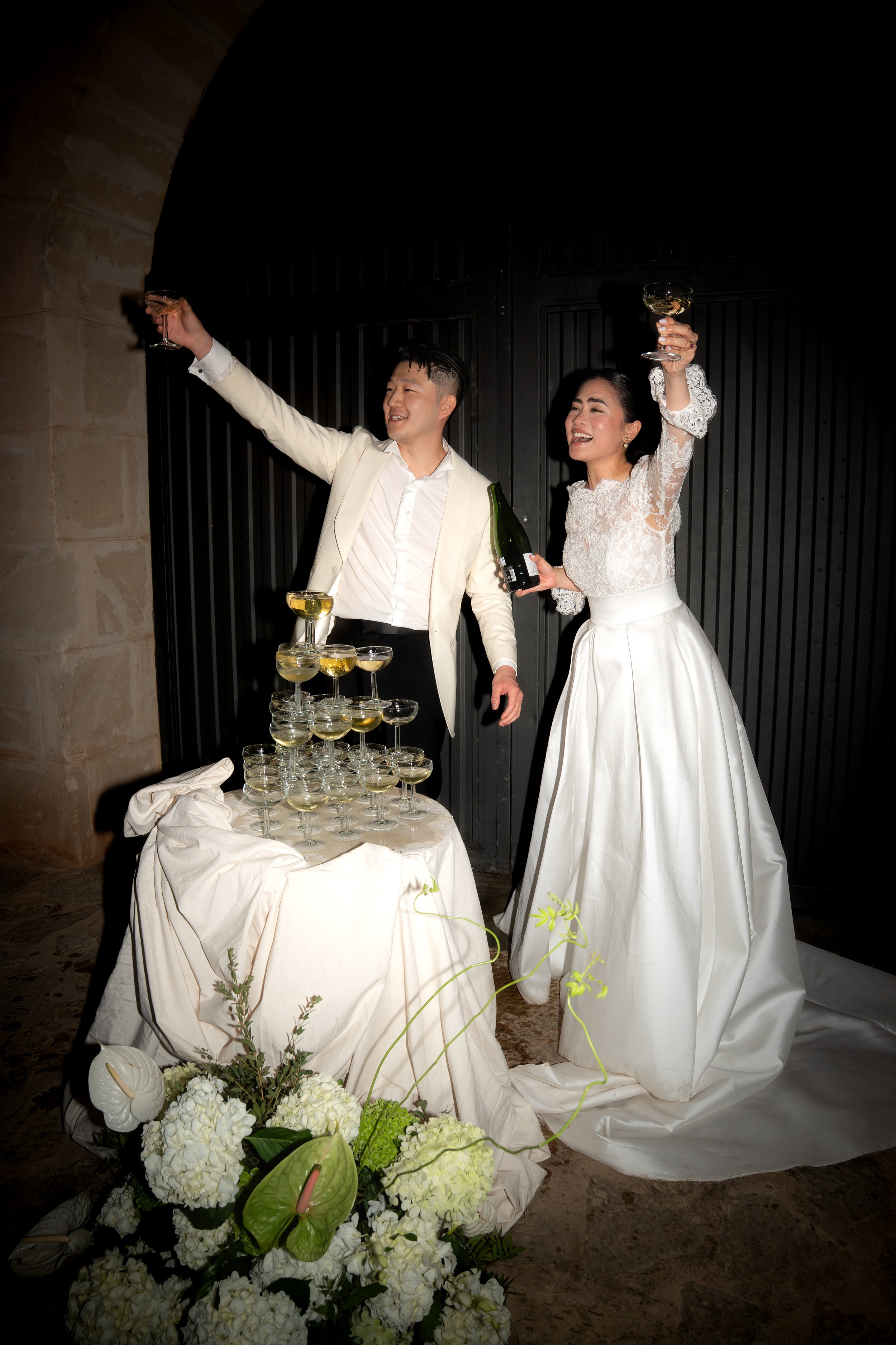 Bride and groom celebrating at their wedding with champagne, standing behind a table with a pyramid of champagne glasses and flowers.