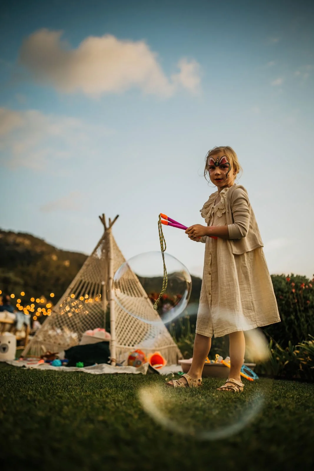 A young girl with butterfly face paint wearing a beige dress and sandals, standing on grass and playing with soap bubbles during sunset. In the background, there is a small teepee tent decorated with lights and various toys, and mountains under a partly cloudy sky.
