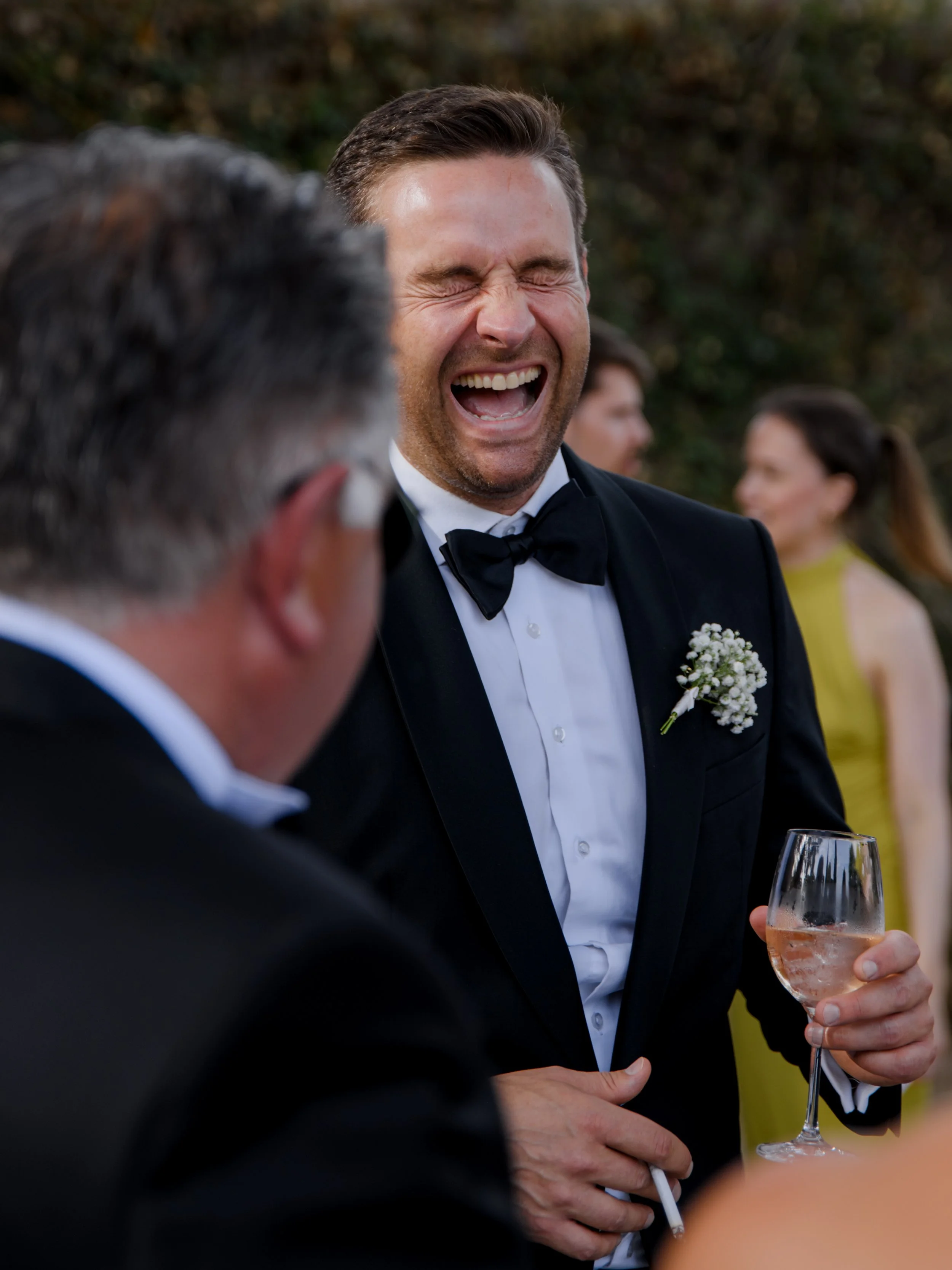 Man in tuxedo laughing and holding a glass of rosé wine at an outdoor event with others in the background.