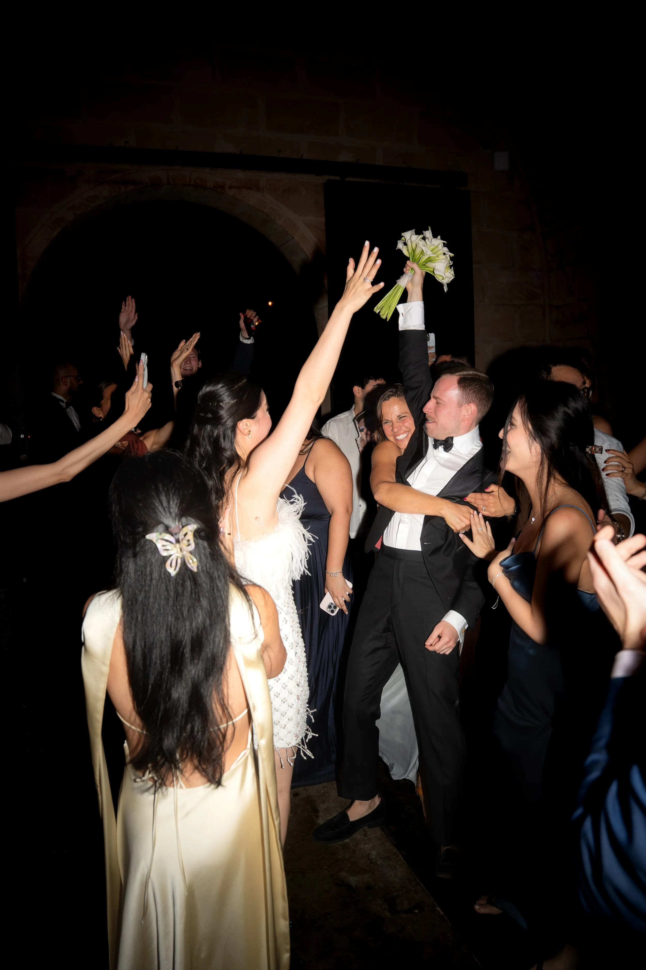 A bride and groom celebrating with friends at a wedding reception, with the groom holding a bouquet of flowers and everyone cheering around them.