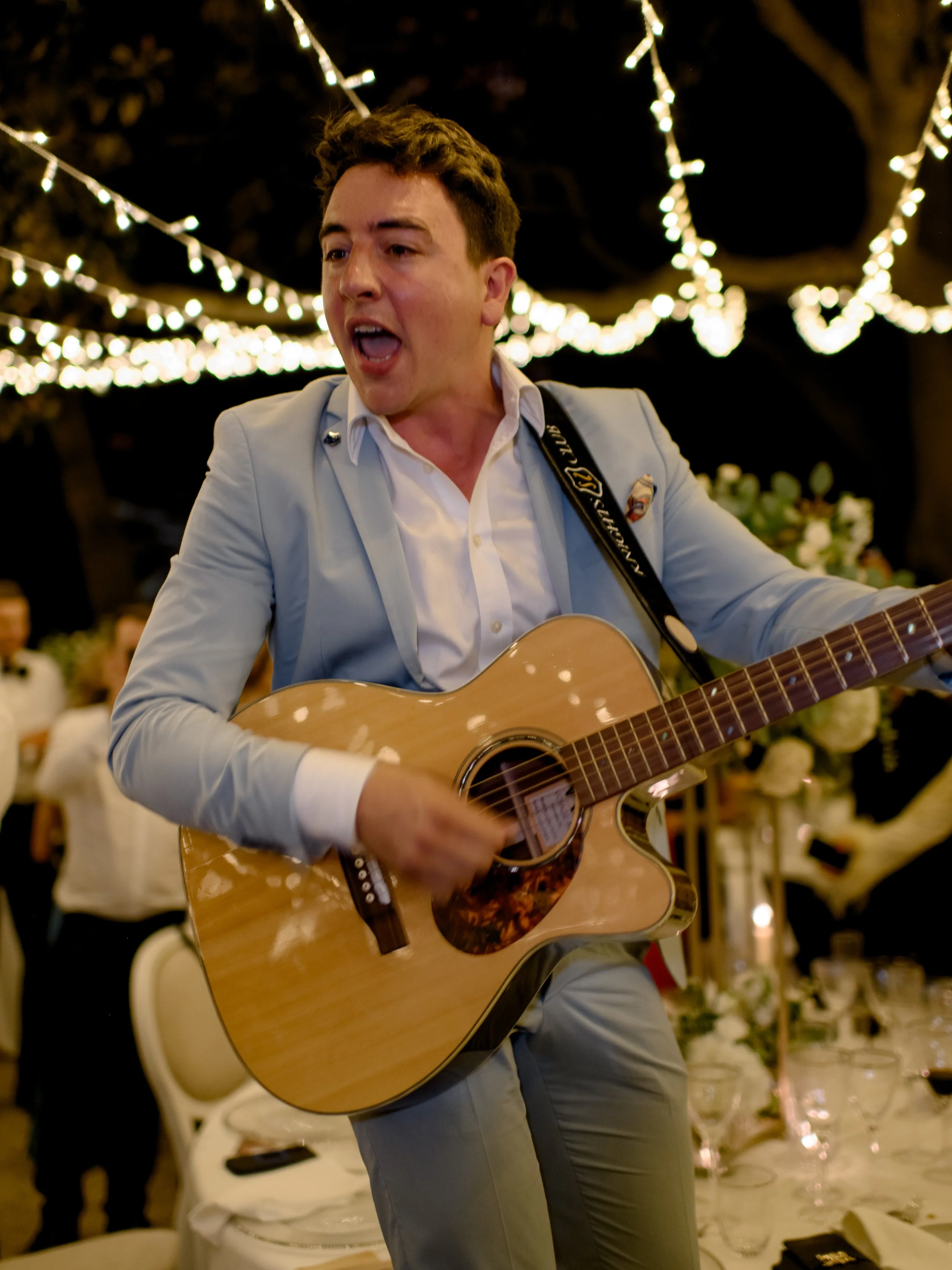 A man in a light-colored suit playing an acoustic guitar at a festive outdoor event with string lights overhead and guests in the background.