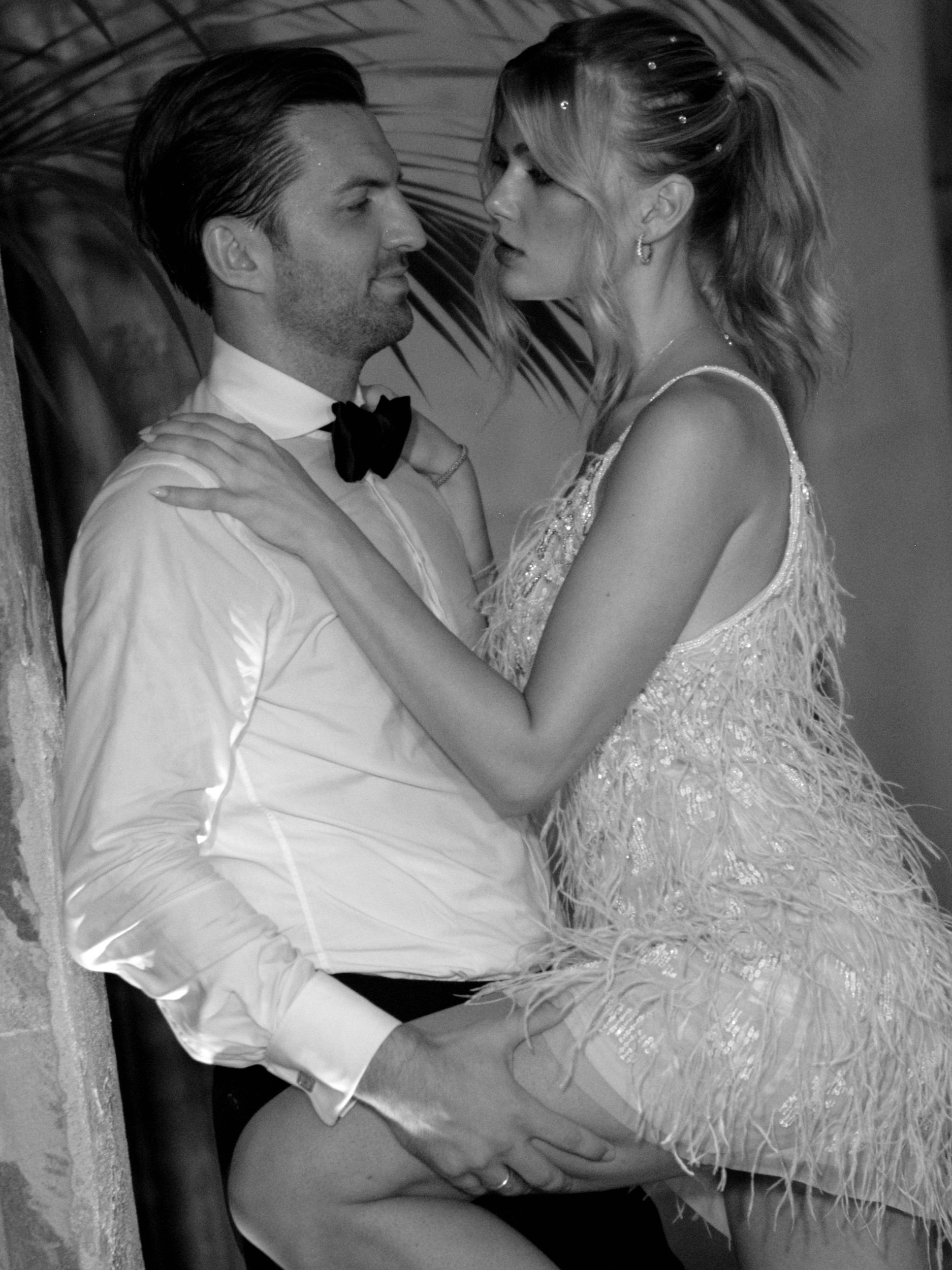 A black and white photo of a man and woman in formal attire. The man is wearing a white shirt and bow tie, while the woman is in a glamorous dress with feathers. They are close, facing each other, with the woman sitting on the man's lap and looking a