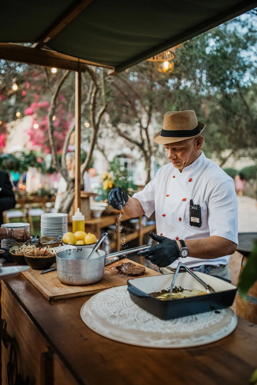 A chef wearing a straw hat, black gloves, and a white apron seasoning a grilled steak on a wooden outdoor kitchen counter, with cooking ingredients and utensils around, under a canopy with string lights in a garden setting.