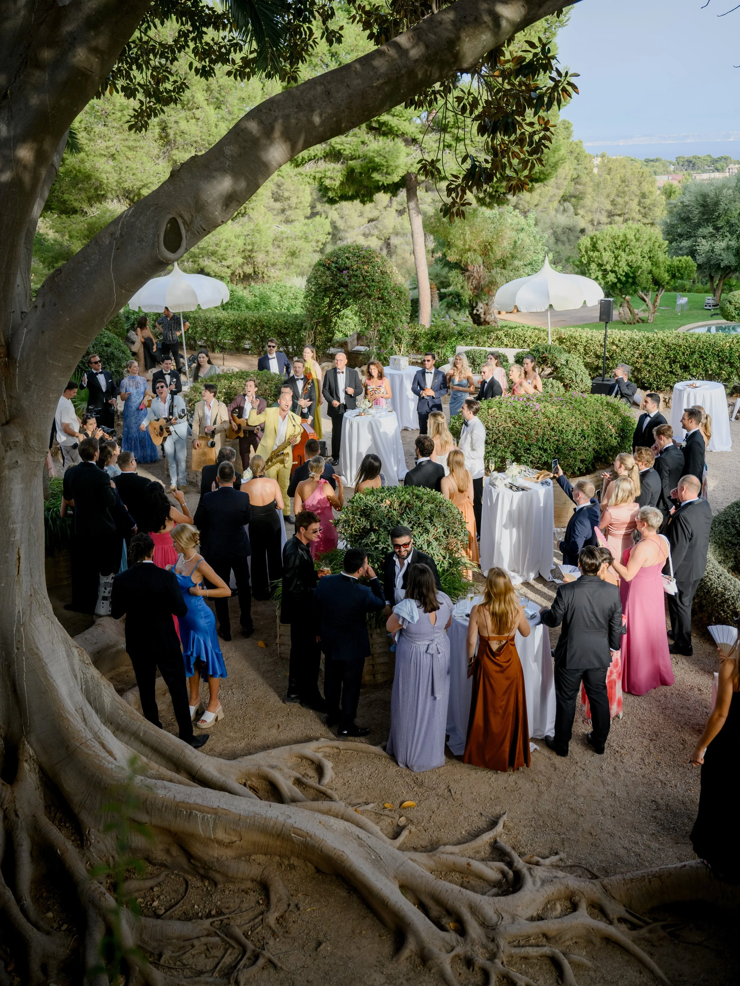 Guests at an outdoor wedding reception with musicians playing guitars, surrounded by trees and decorated with umbrellas.
