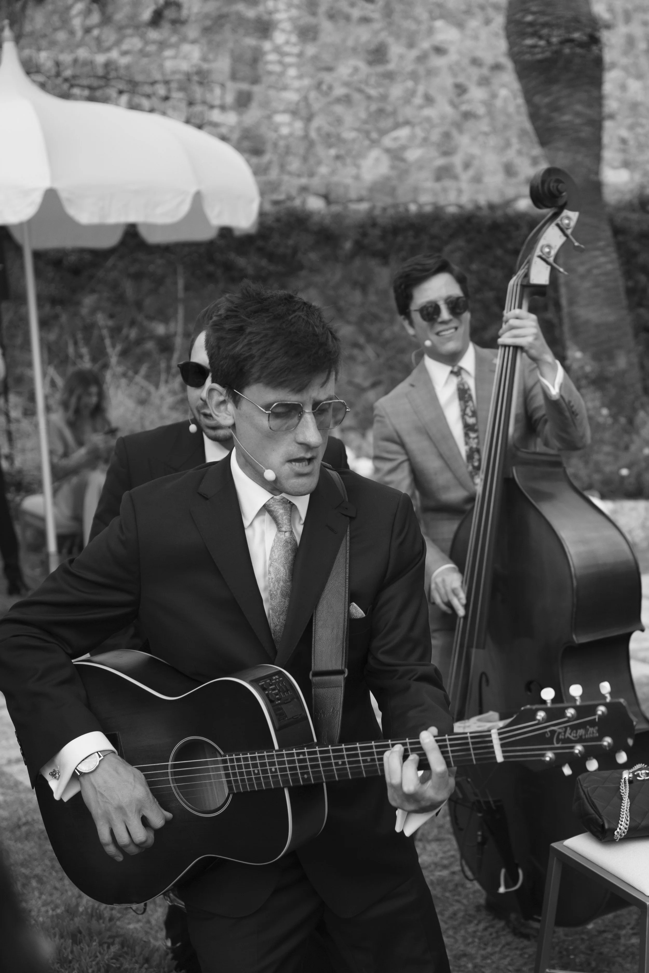 A man in a suit and sunglasses plays an acoustic guitar while a man in a suit and sunglasses plays a double bass in an outdoor setting.