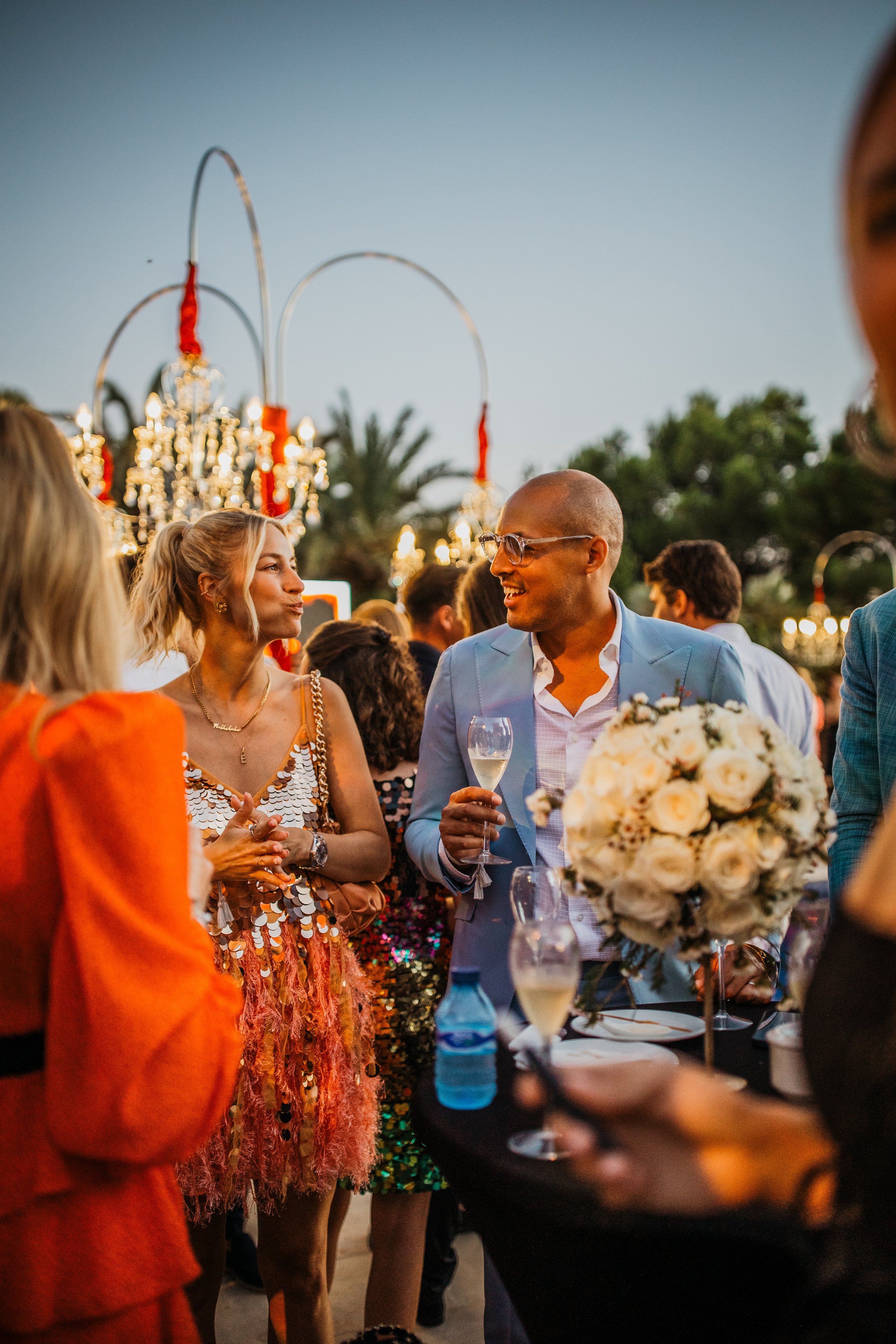 People talking and celebrating outdoors at a wedding reception during sunset, with floral arrangements and chandeliers in the background.