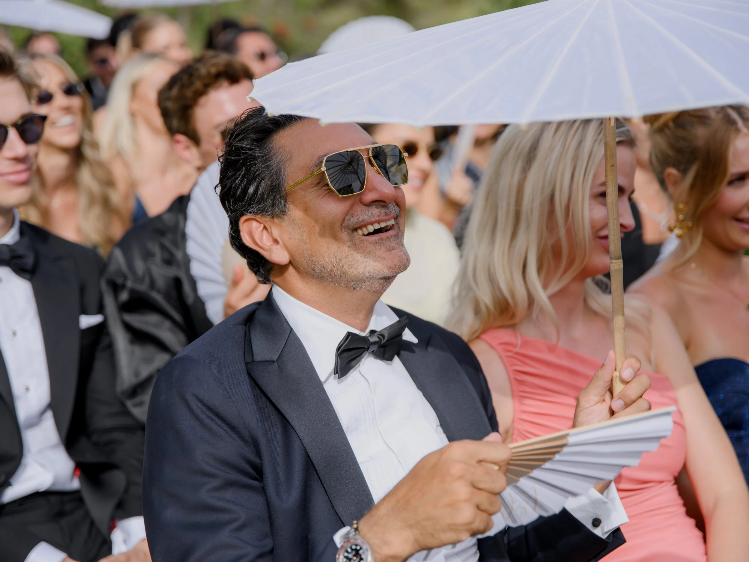 A group of well-dressed people at an outdoor event, with a man in the foreground wearing sunglasses, a tuxedo, and holding a fan while smiling. A woman behind him holds a white umbrella over her head.