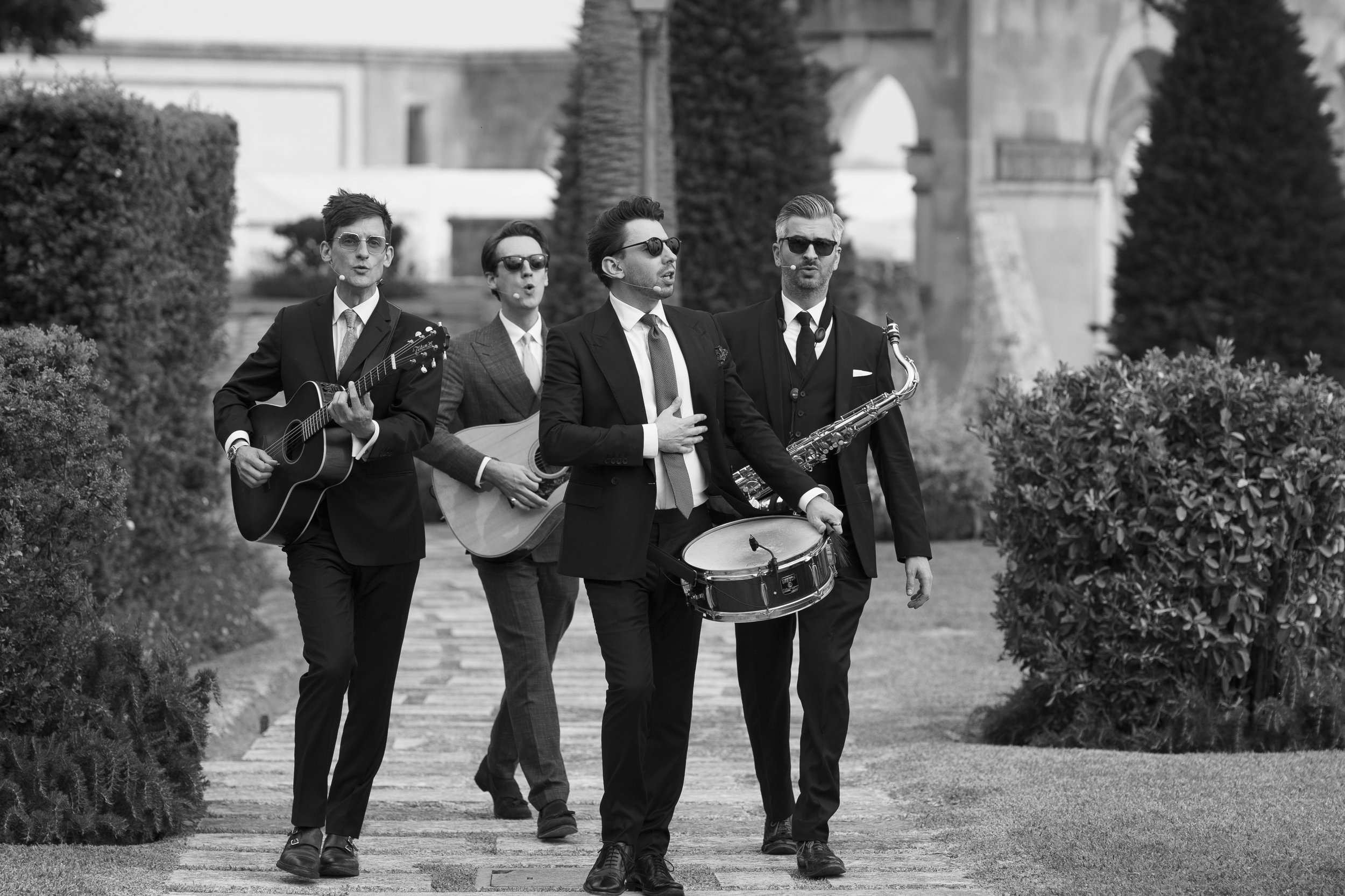 A black and white photo of a five-piece jazz band walking outdoors on a pathway with trees and a building in the background. The band members are dressed in formal suits and are playing various instruments including guitar, drums, and saxophone.