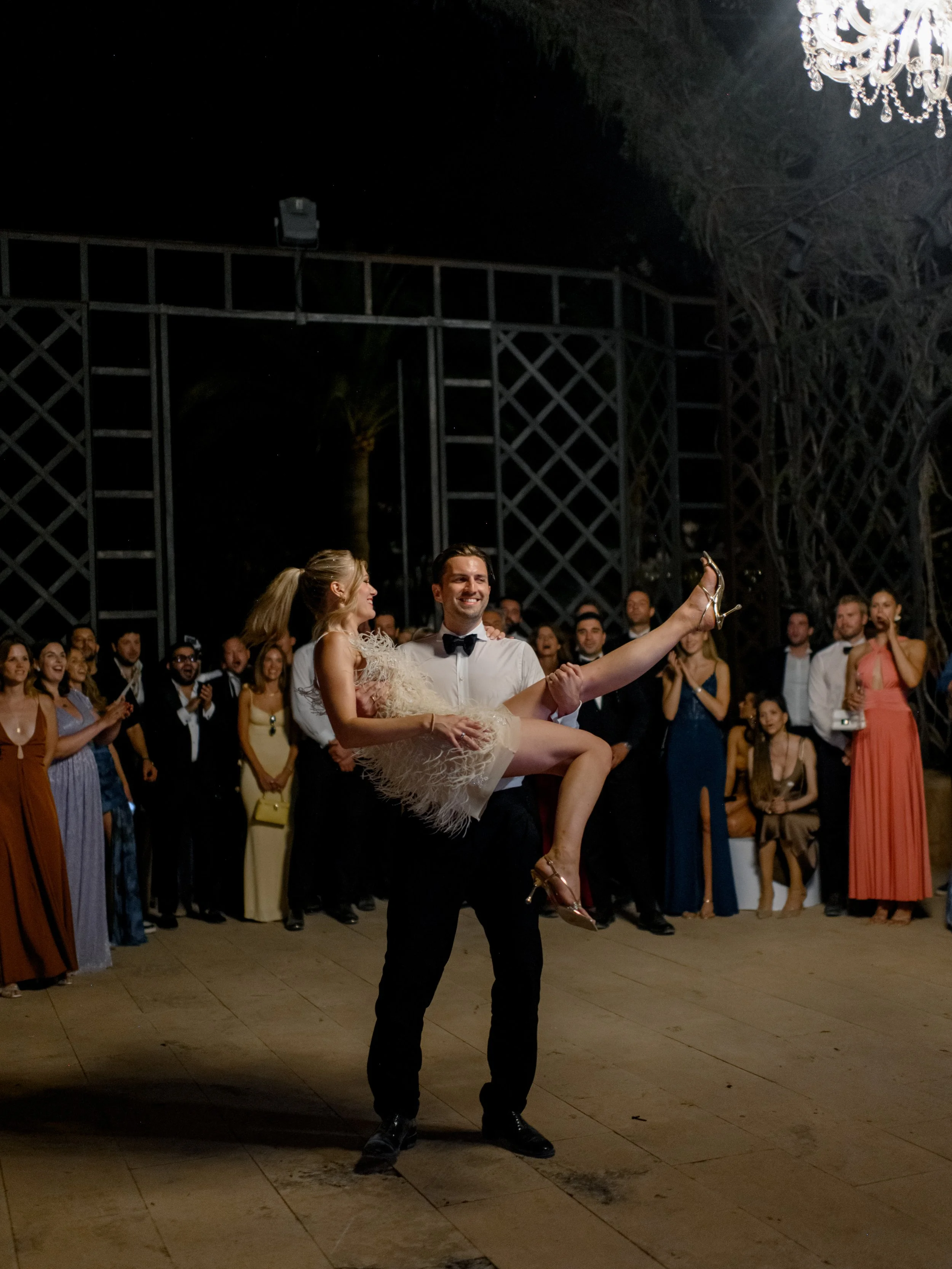 A groom dances with a bride in a white feathered dress at a nighttime wedding reception, surrounded by well-dressed guests clapping and smiling.