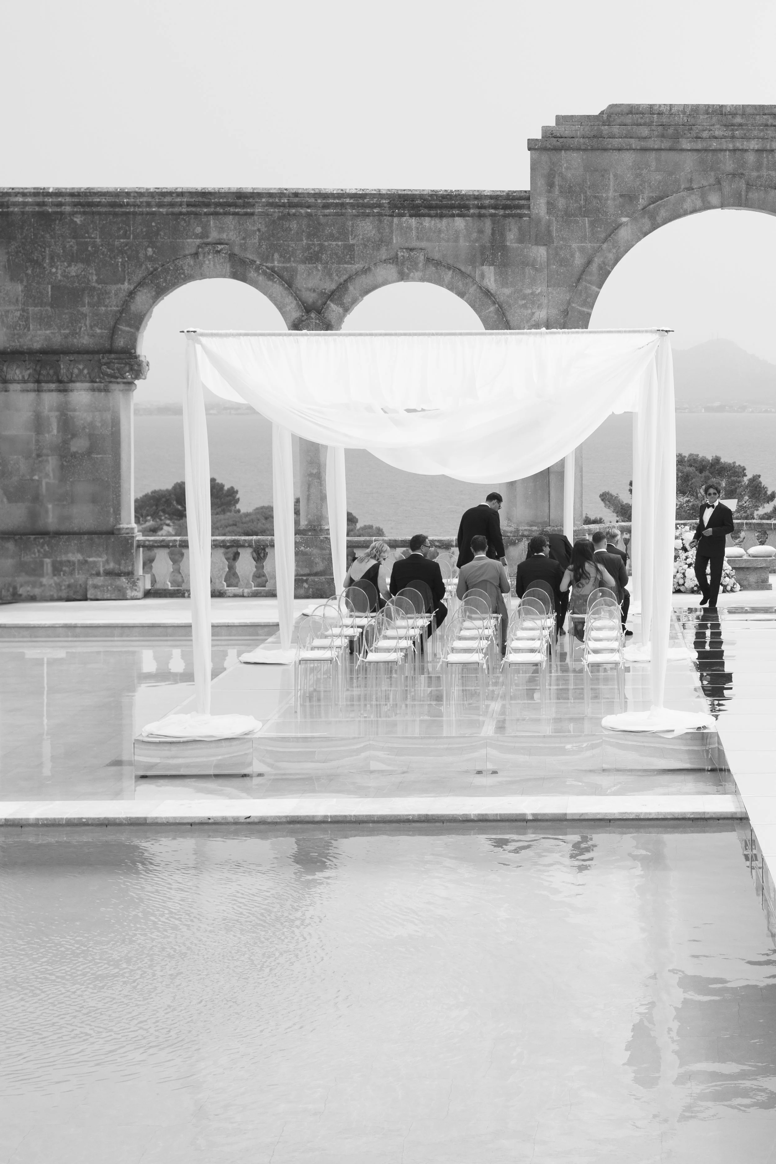 Wedding ceremony setup outdoors on a terrace with a view of the water and mountains in the background. There is a canopy with draped fabric, chairs arranged in rows, and people dressed formally, possibly preparing for the ceremony.