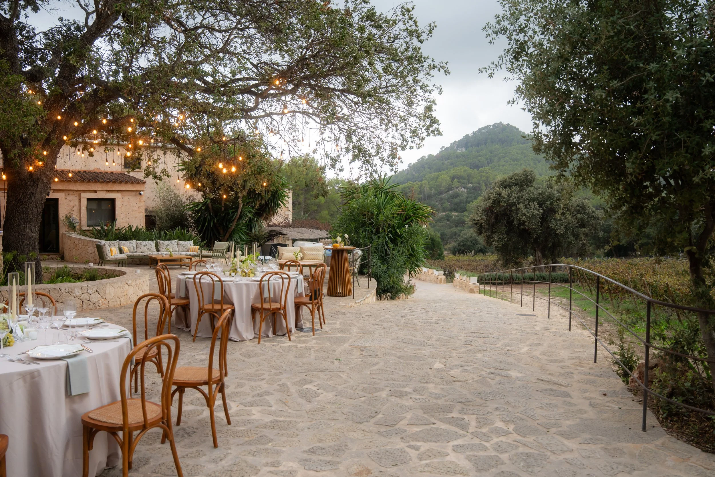 Outdoor dining setup with round tables and chairs, decorated with white tablecloths, under a large tree with string lights, in a scenic mountainous landscape with greenery and stone pathways.