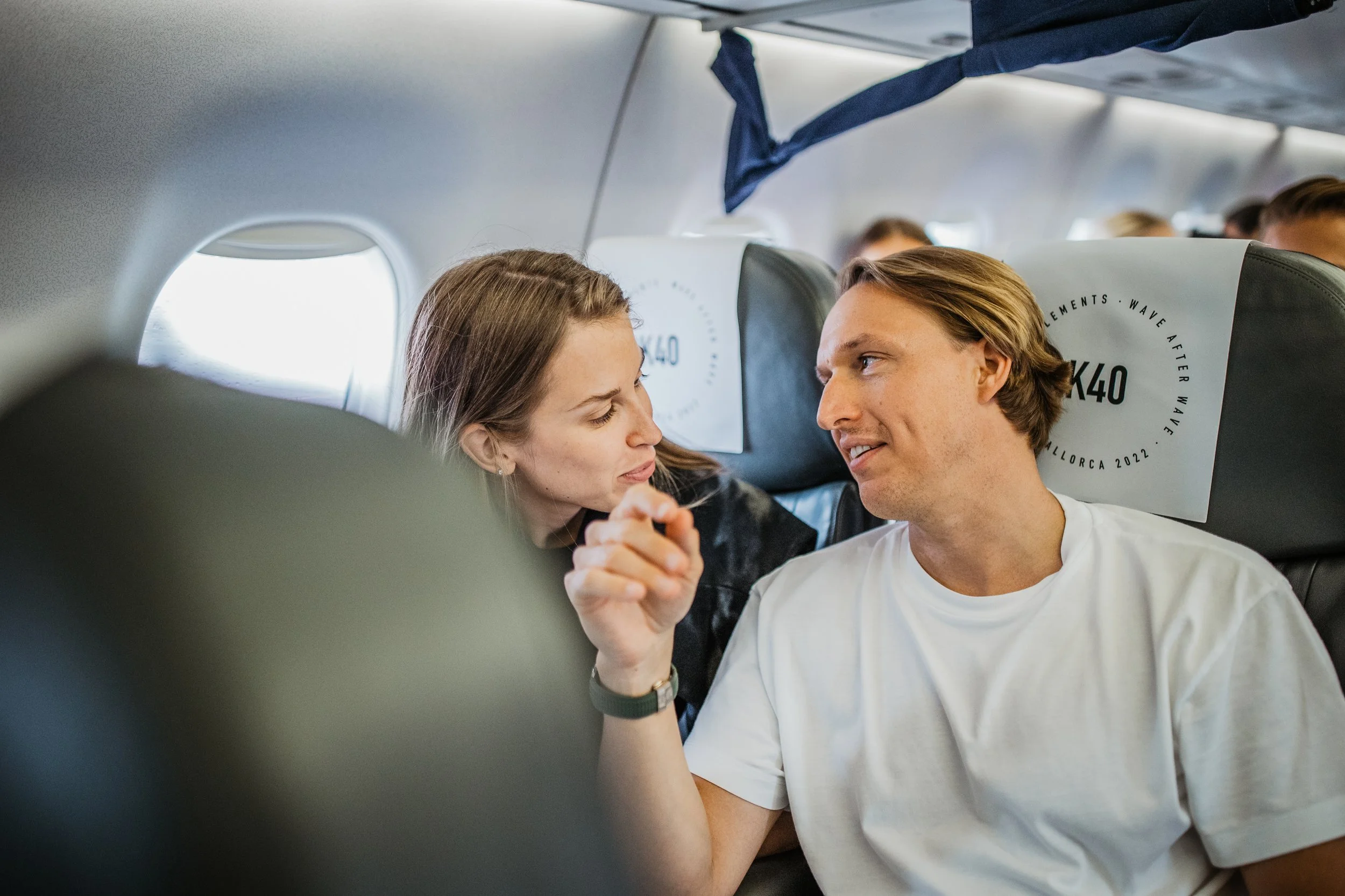 A man and woman sitting next to each other on an airplane, engaged in conversation with the woman leaning in towards the man at their seats.