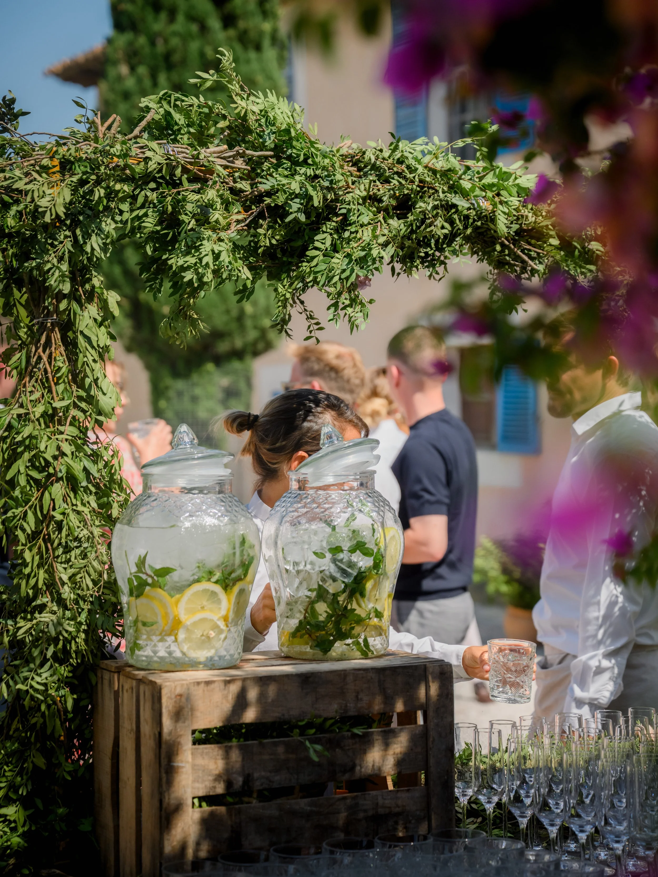 People at an outdoor event with glass dispensers filled with lemonade and mint leaves, surrounded by greenery and colorful flowers, engaging in conversation under a leafy arch.