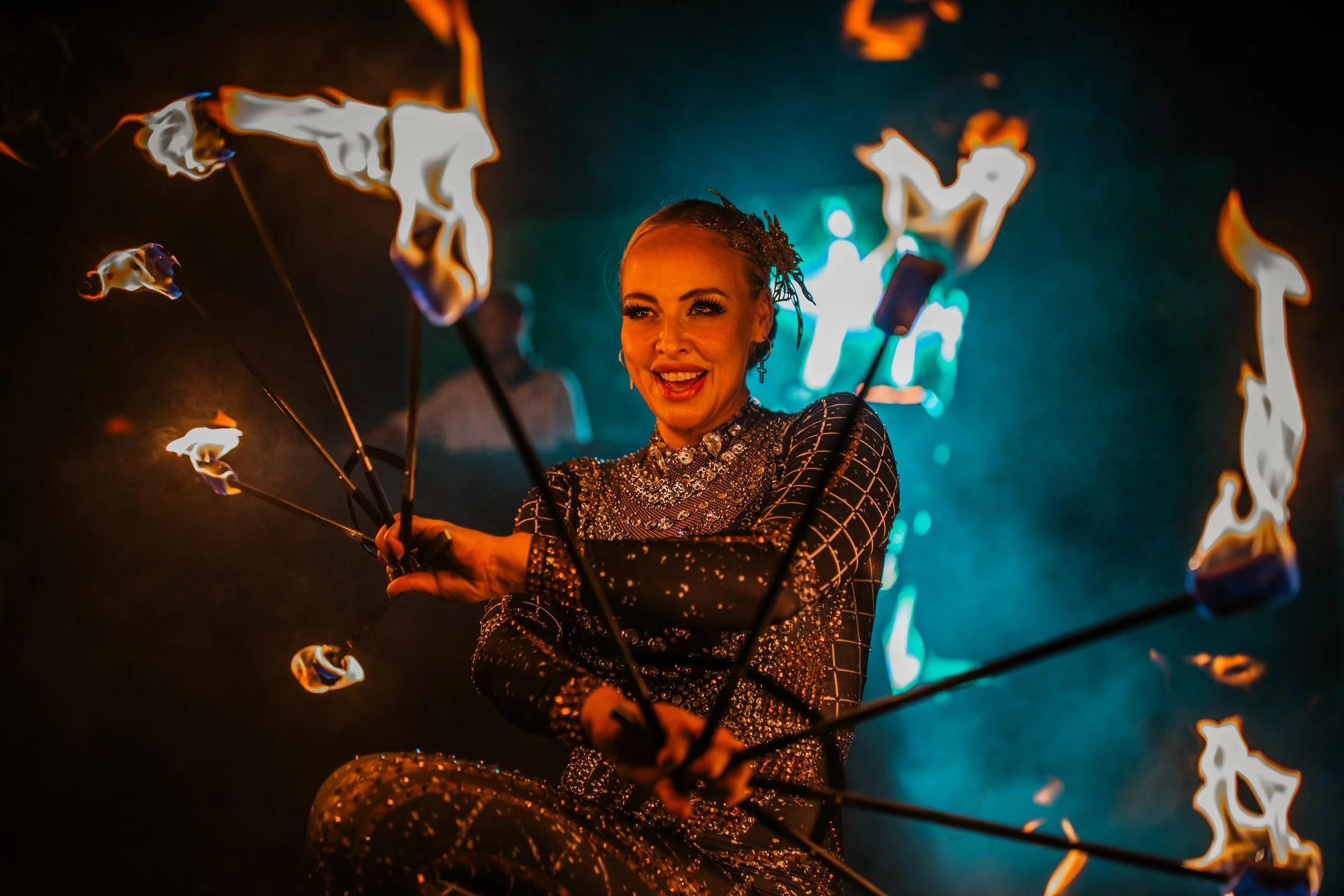 A female performer spinning flaming poi sticks in a dimly lit environment with colorful lighting in the background, smiling.