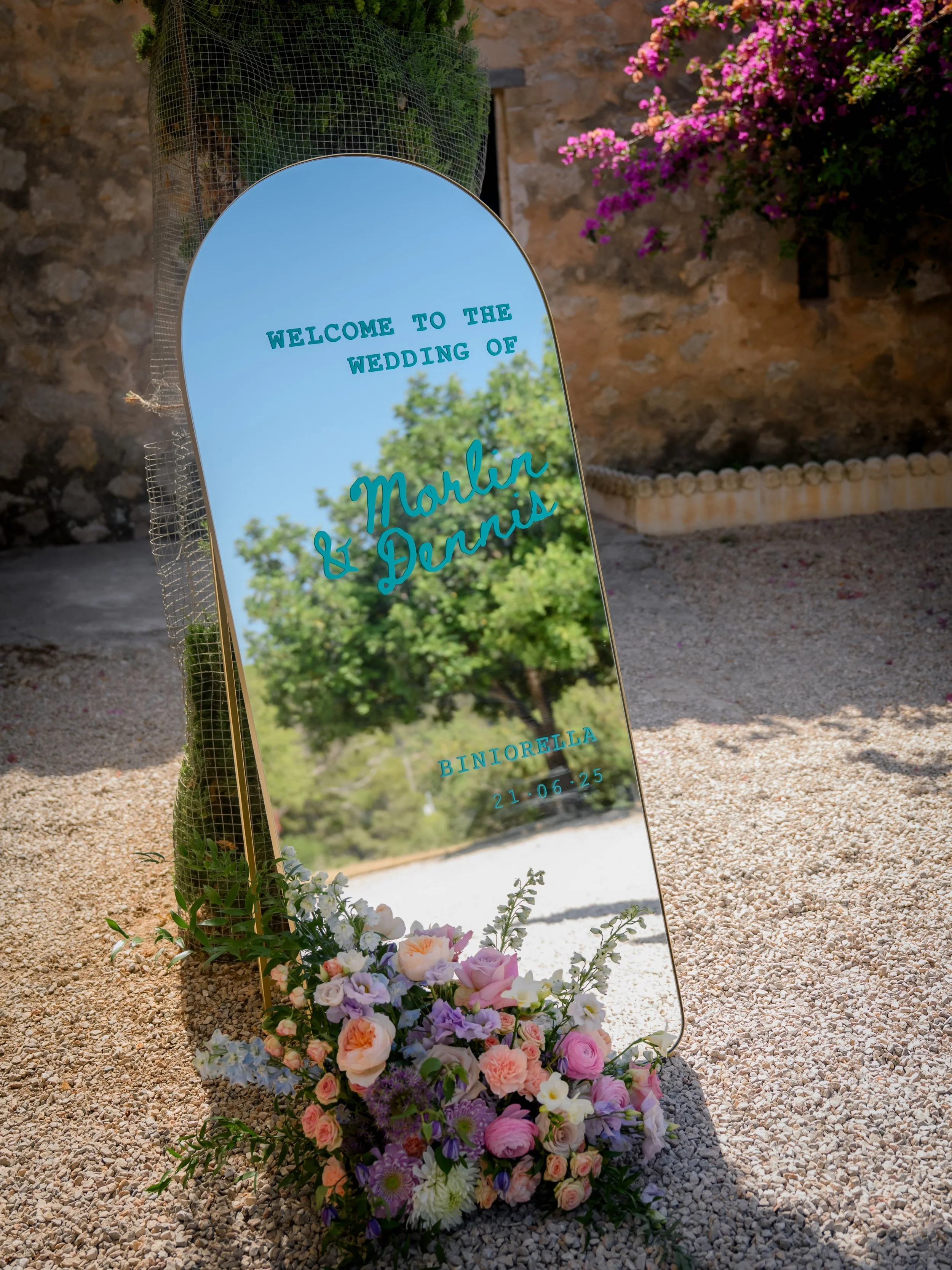 A decorative mirror wedding sign with blue text welcomes guests to the wedding of Marlin and Danni. The sign is adorned with a bouquet of pink, purple, and white flowers at its base and is set on sandy ground with a stone wall and pink flowering shru