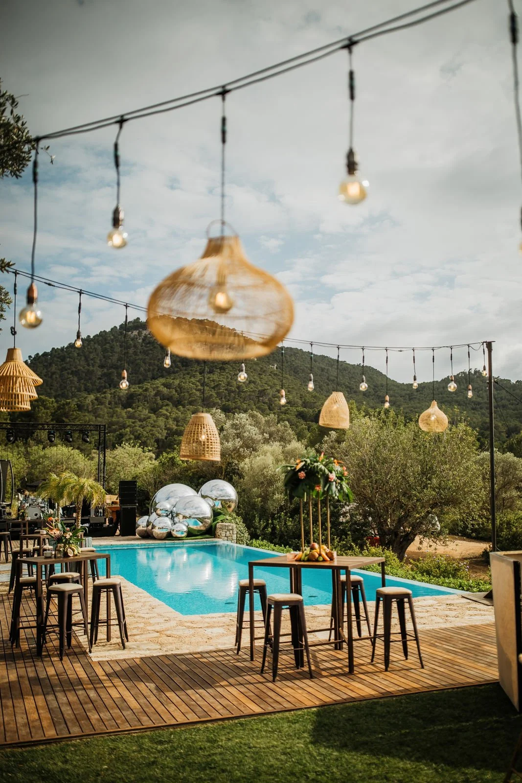 Outdoor poolside setting with hanging string lights, wicker and glass ornaments, a high table with chairs, a floral centerpiece, and lush green trees and hills in the background.