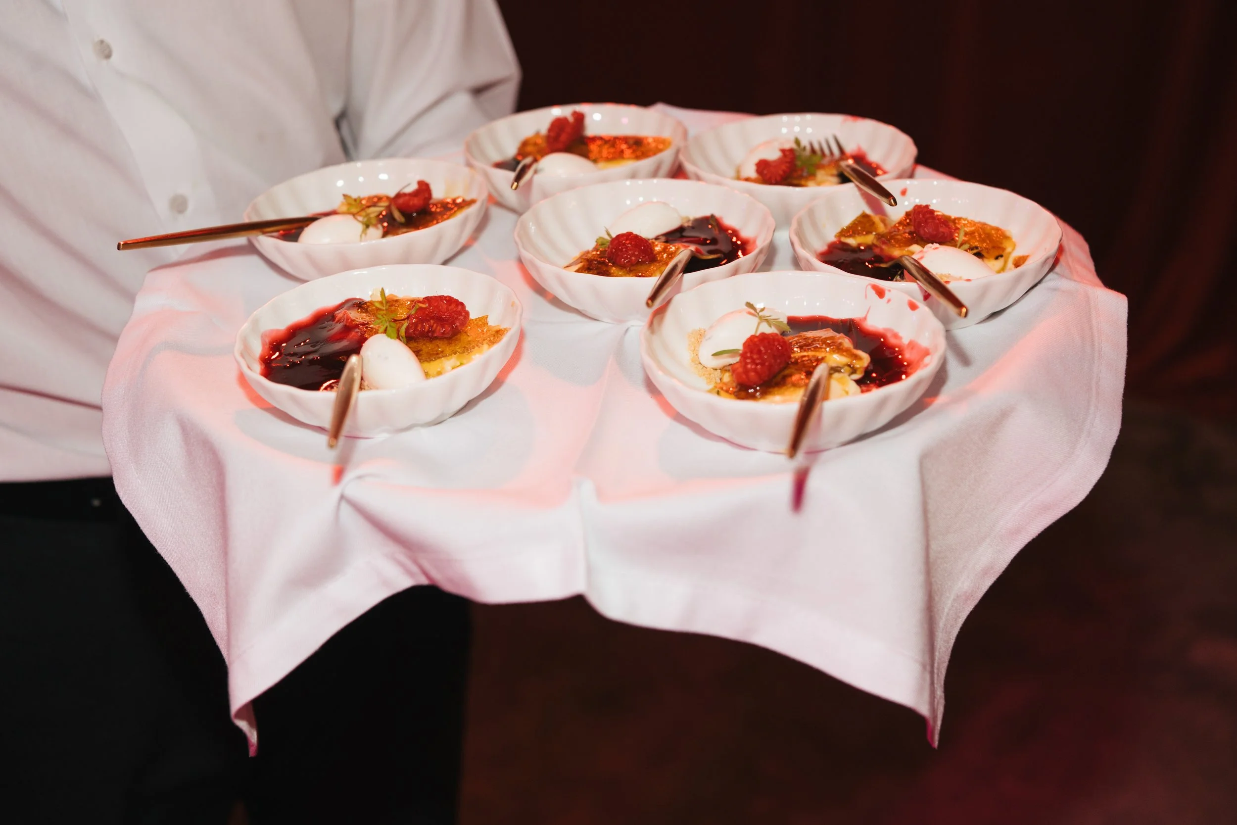 Person holding a tray with nine small dessert bowls containing cream, raspberry sauce, and garnishes.