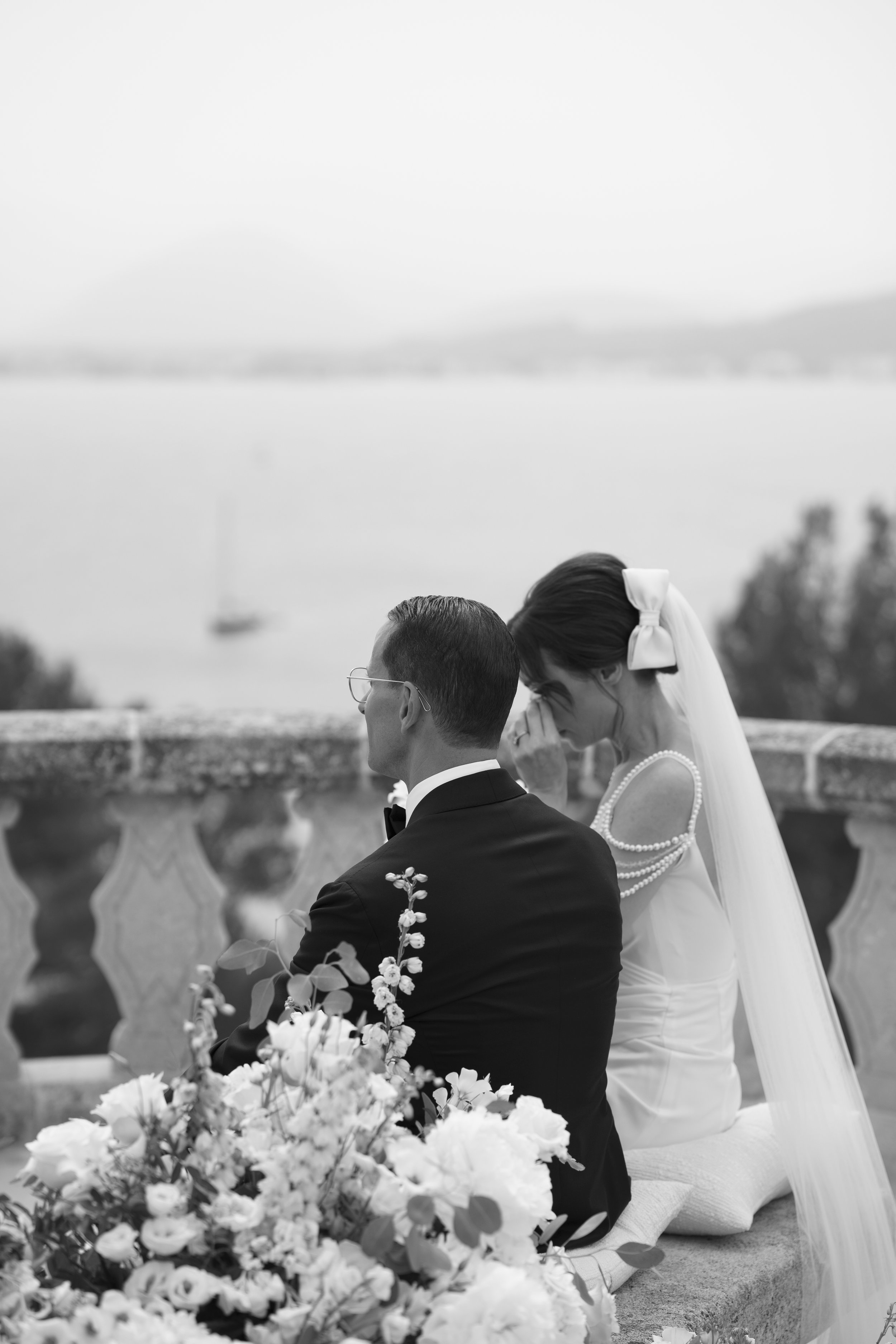 A black and white photo of a bride and groom sitting on a stone bench outdoors, with the groom facing away and the bride leaning forward with her hand near her face, overlooking a body of water with a sailboat in the distance.