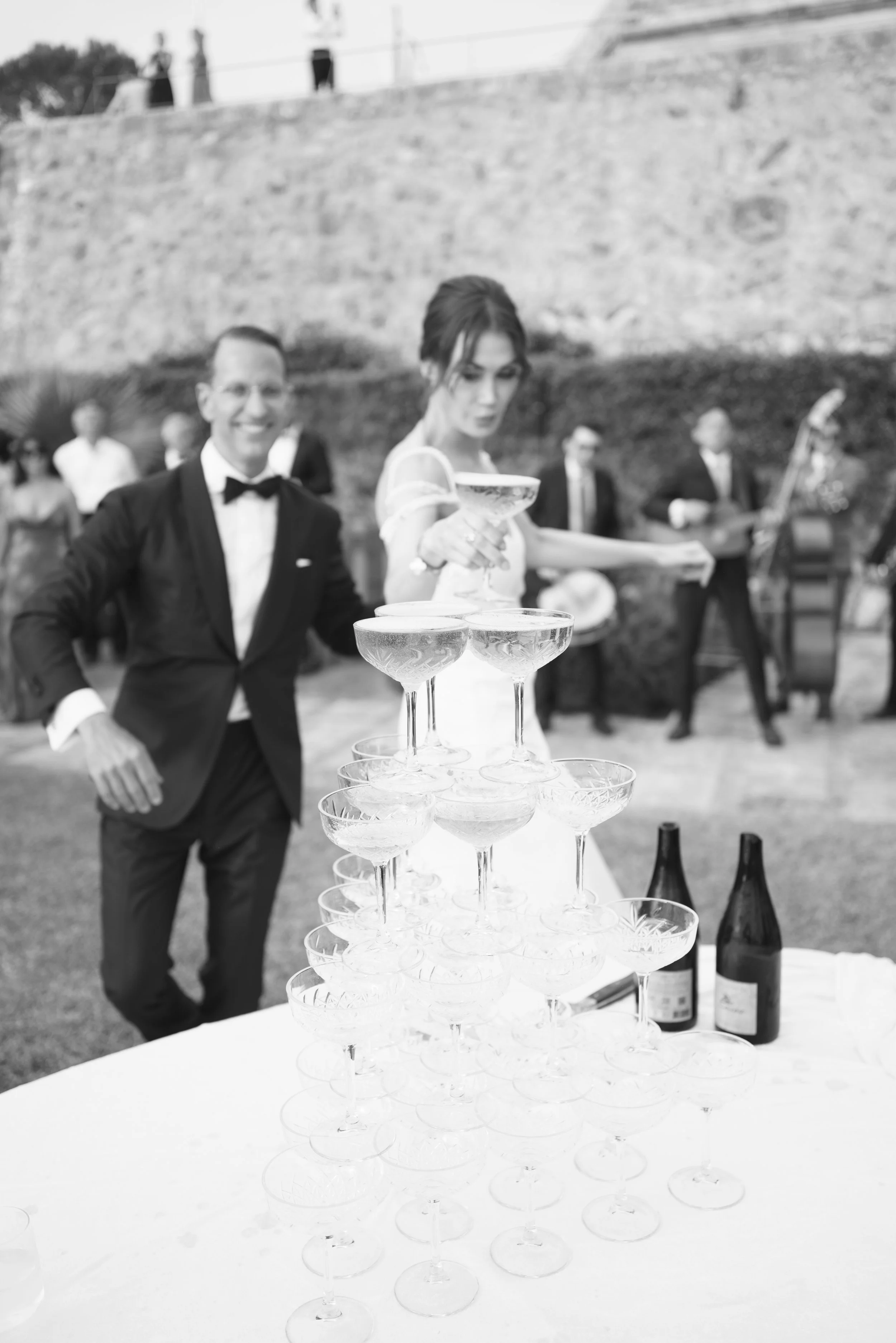 Black and white photo of a wedding celebration with a champagne tower, a bride in a wedding dress, a groom in a tuxedo, and a band performing in the background.