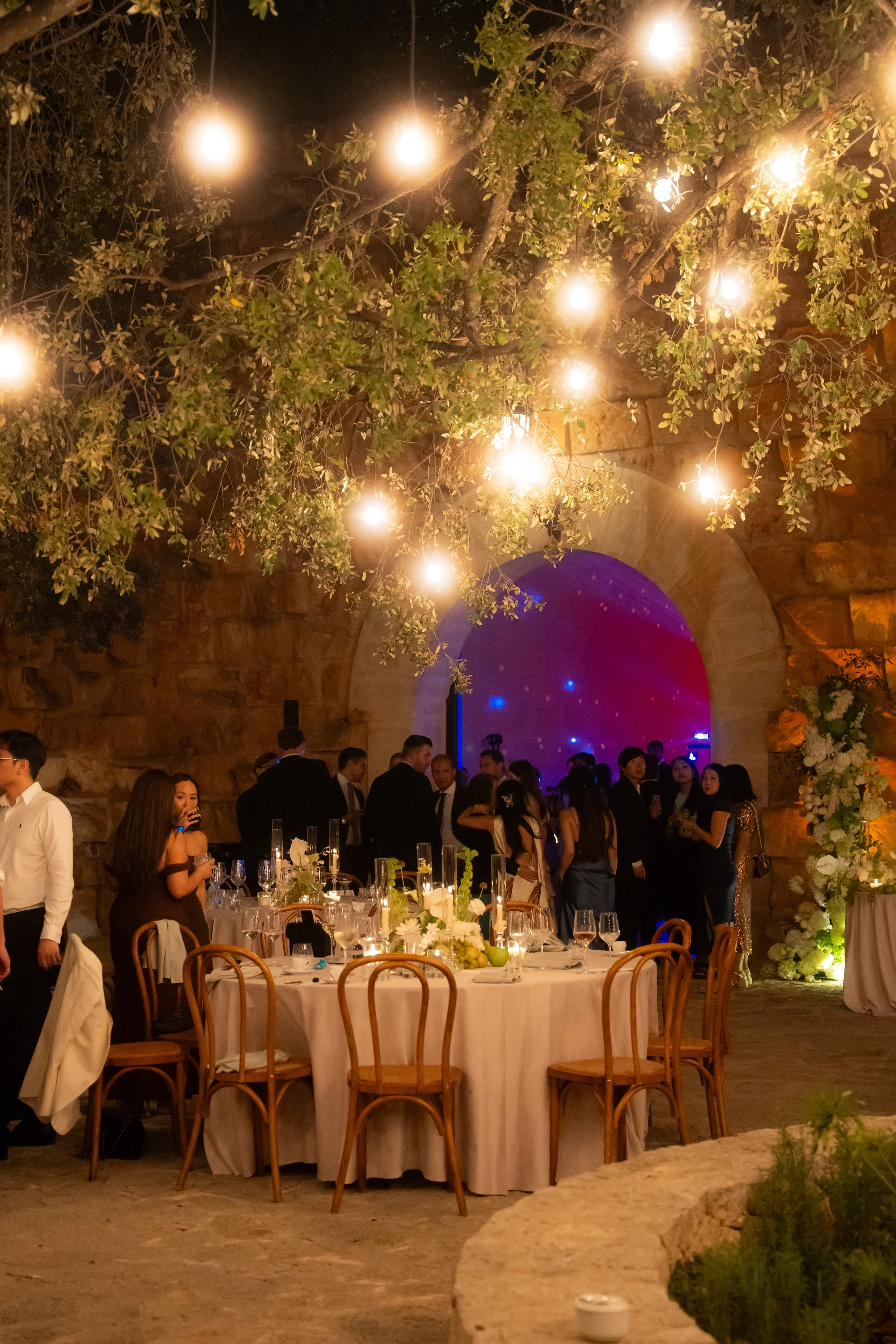 A wedding or formal event inside a decorated venue with warm lighting, round tables with white tablecloths and floral centerpieces, and guests dressed in formal attire socializing under hanging lights and a large stone archway.