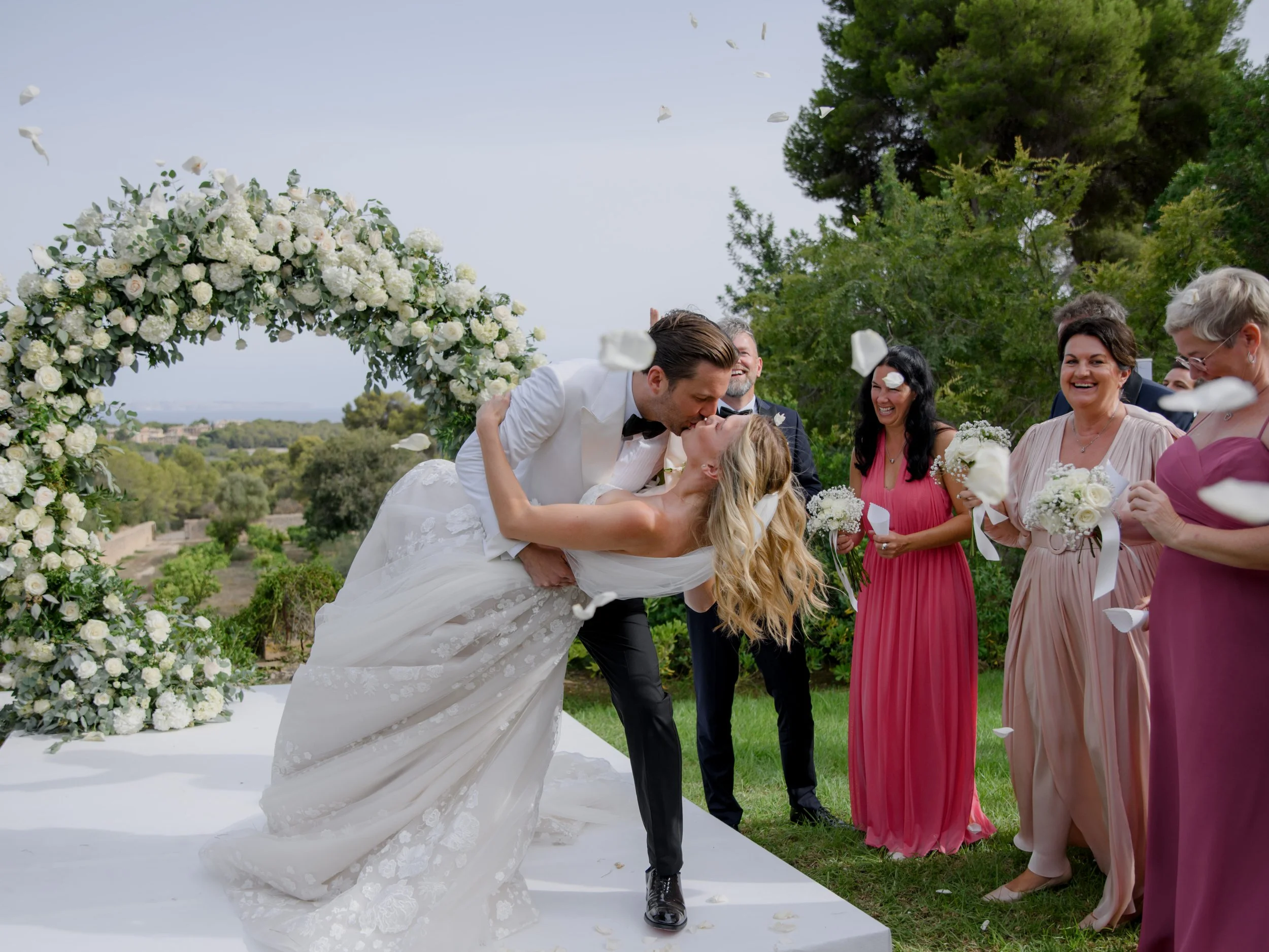 A newlywed couple sharing a kiss during their outdoor wedding ceremony with a floral arch and bridesmaids in colorful dresses.