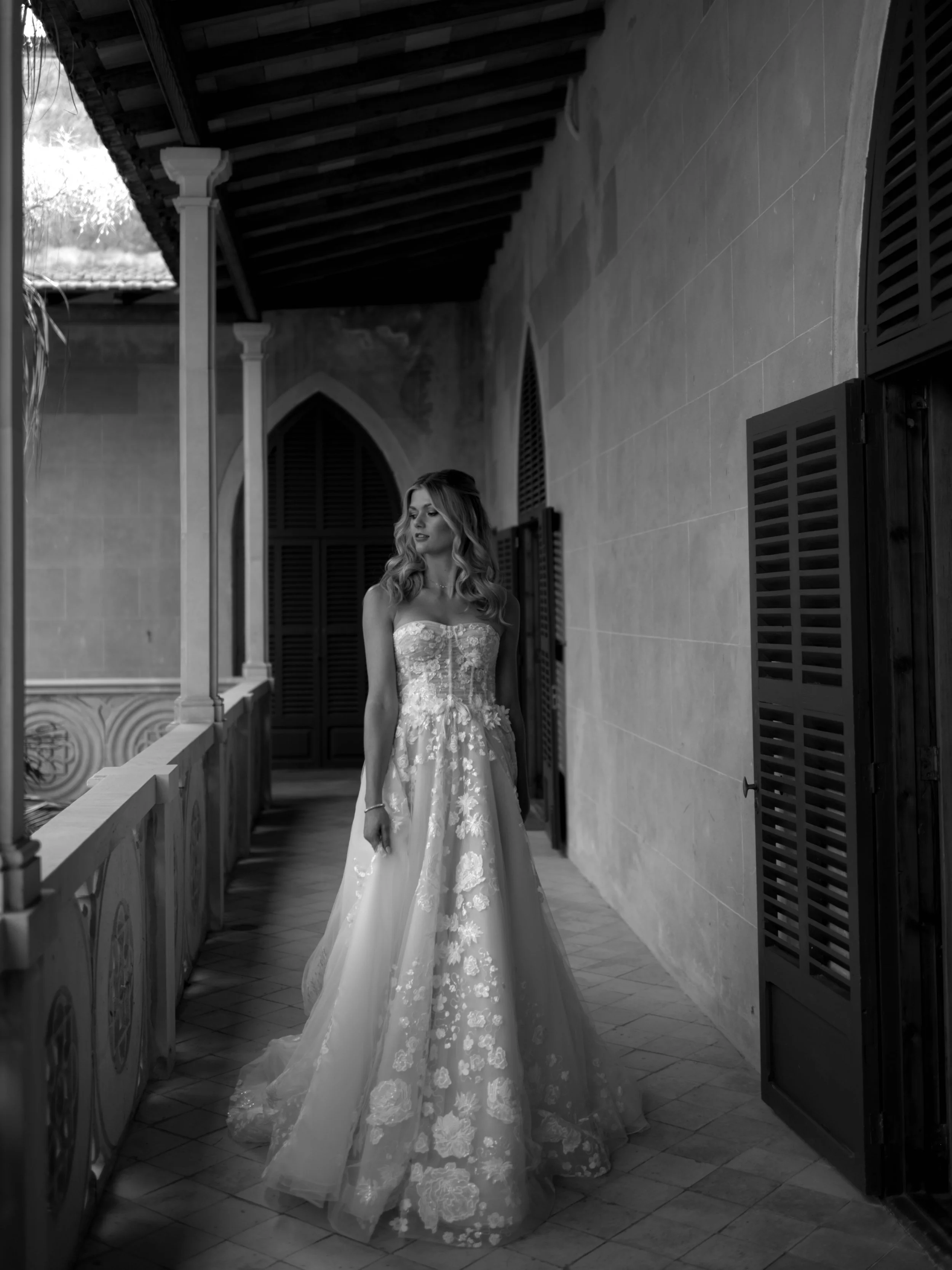A woman in a wedding dress standing on a balcony with wooden shutters on the window and a tiled floor, looking slightly to the side.