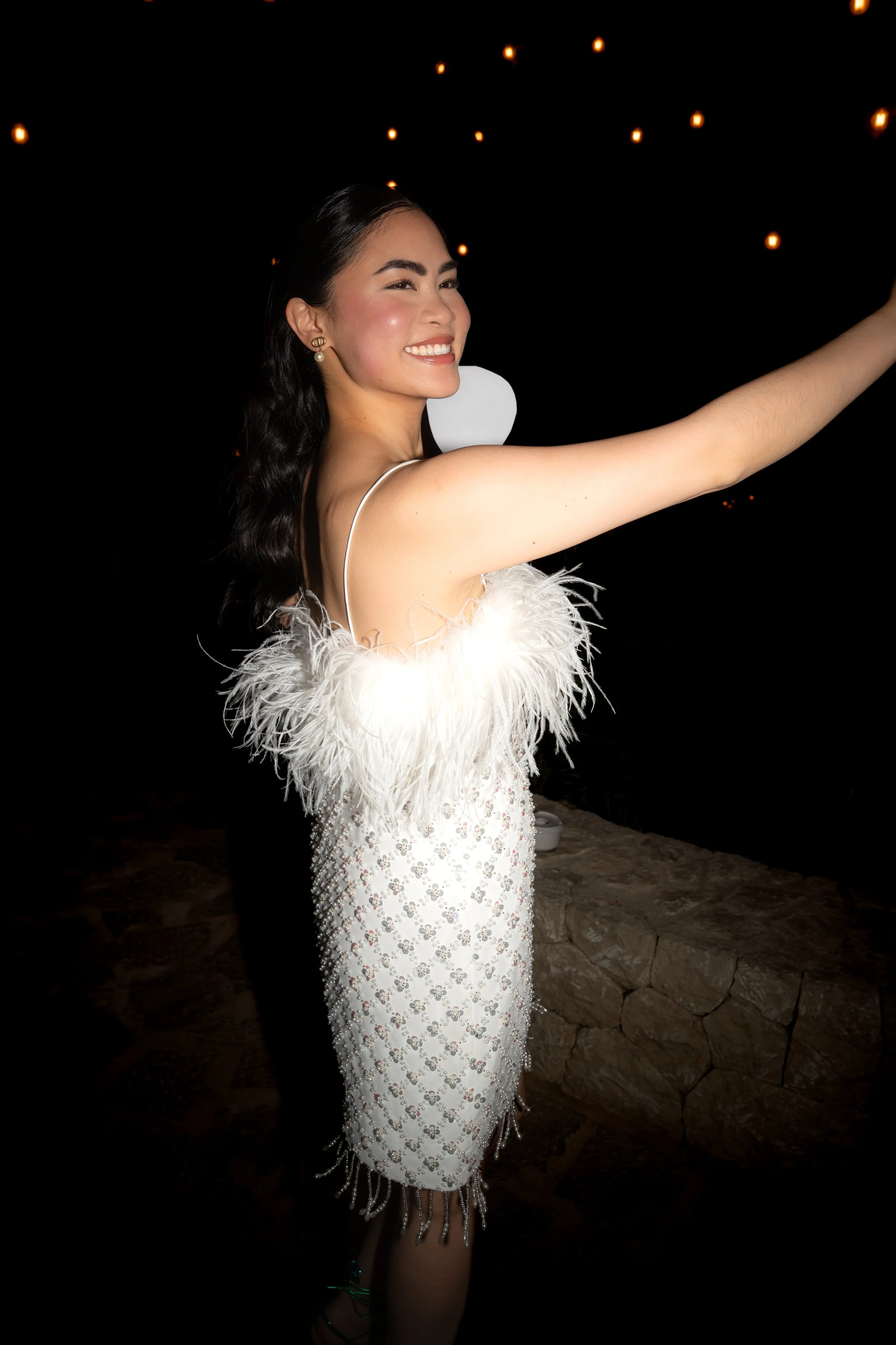 A woman in a glamorous white dress with feather accents and embellishments, smiling and taking a selfie outdoors at night with string lights overhead.