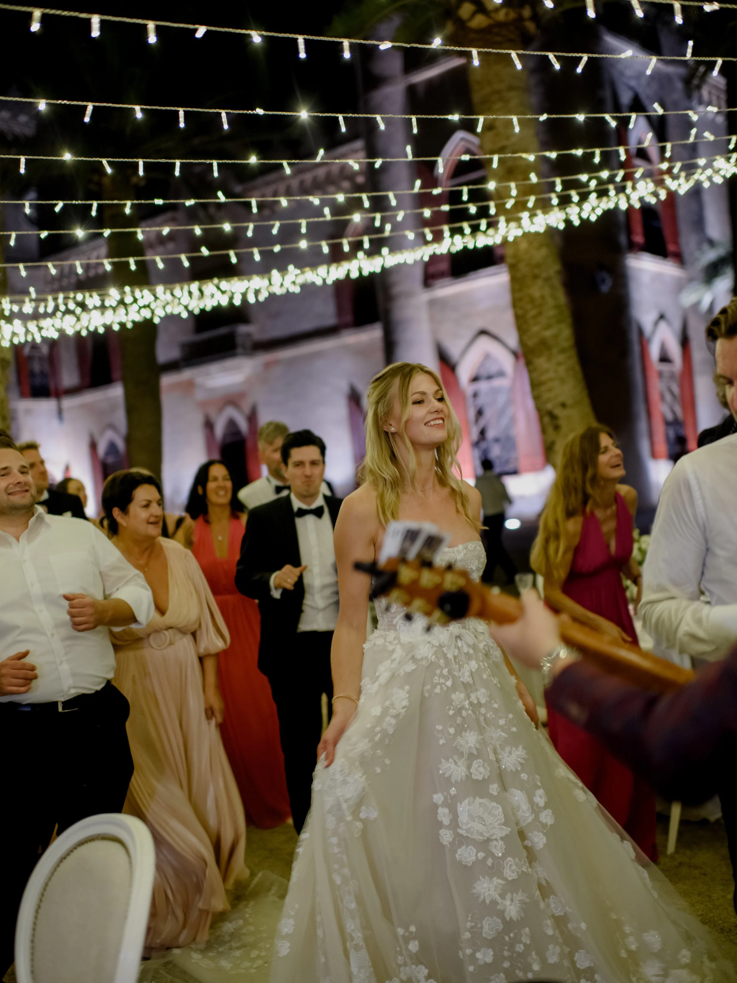 A bride in a white wedding dress smiling at a wedding reception, surrounded by guests dancing and celebrating under string lights at night.