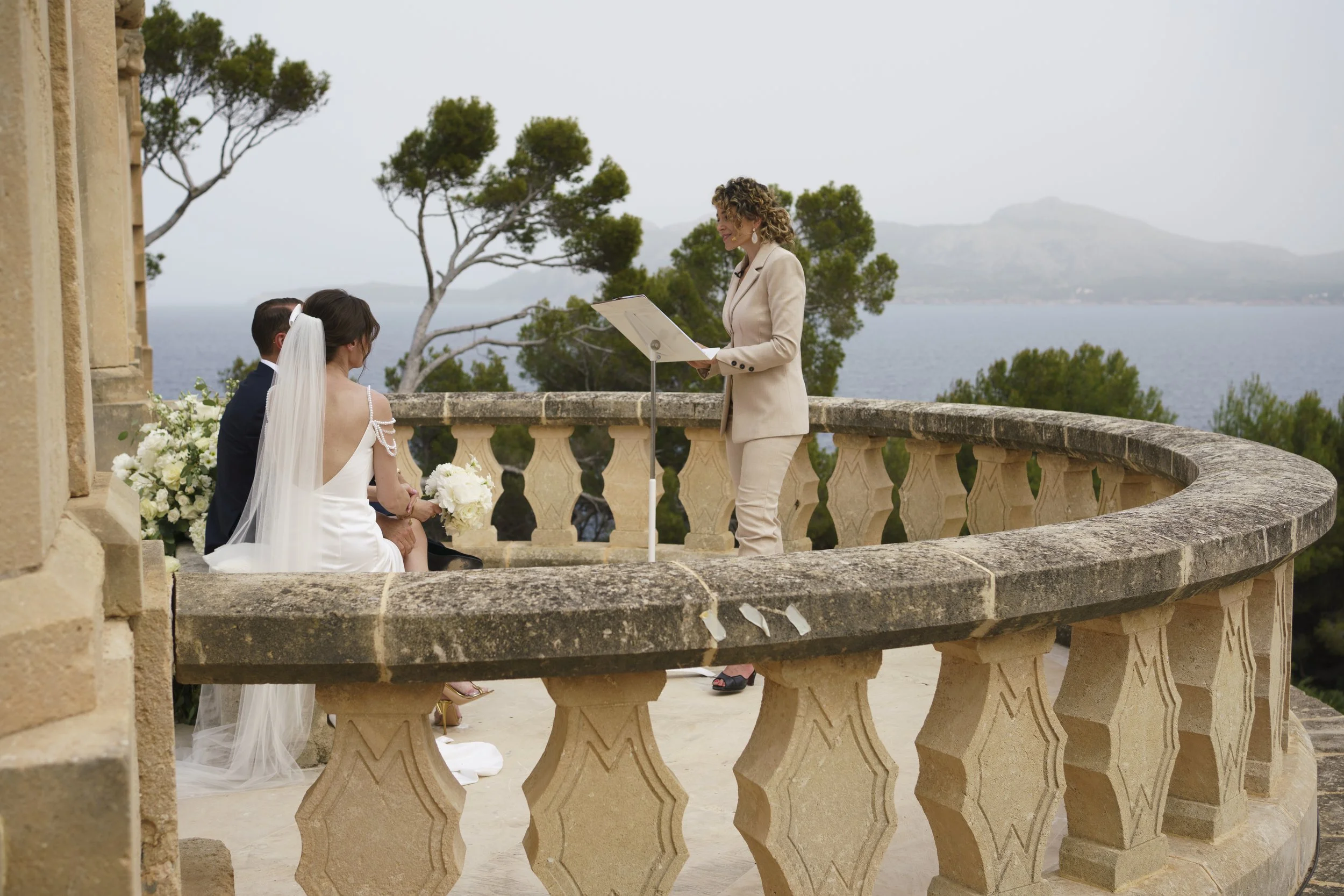 A wedding ceremony taking place on a balcony overlooking a lake and trees, with an officiant reading from a document to a bride and groom.