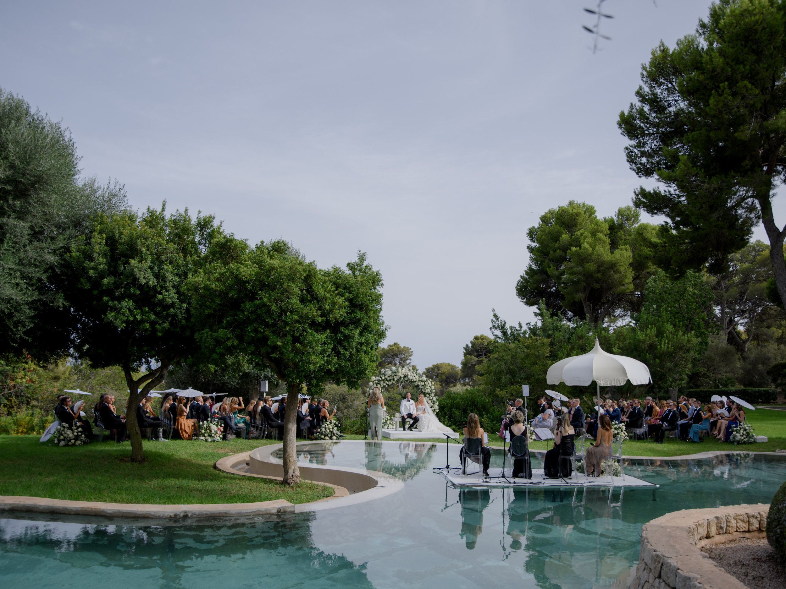 Outdoor wedding ceremony by a pool with seated guests, officiant, and bride and groom, surrounded by trees and greenery.